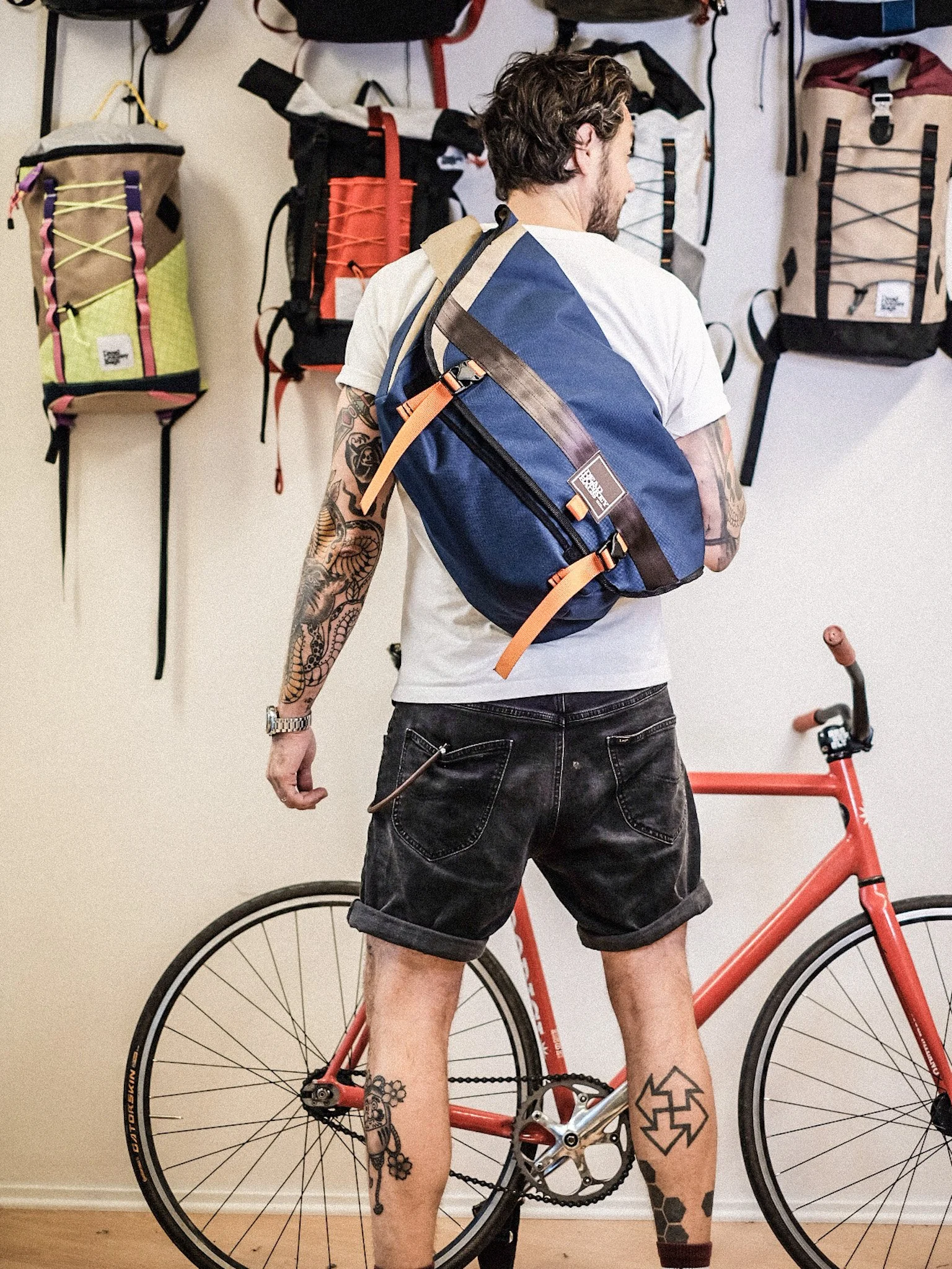 A man with tattoos on his right arm and legs stands in a bike shop, looking at a row of backpacks hanging on the wall. A red bicycle is positioned nearby.