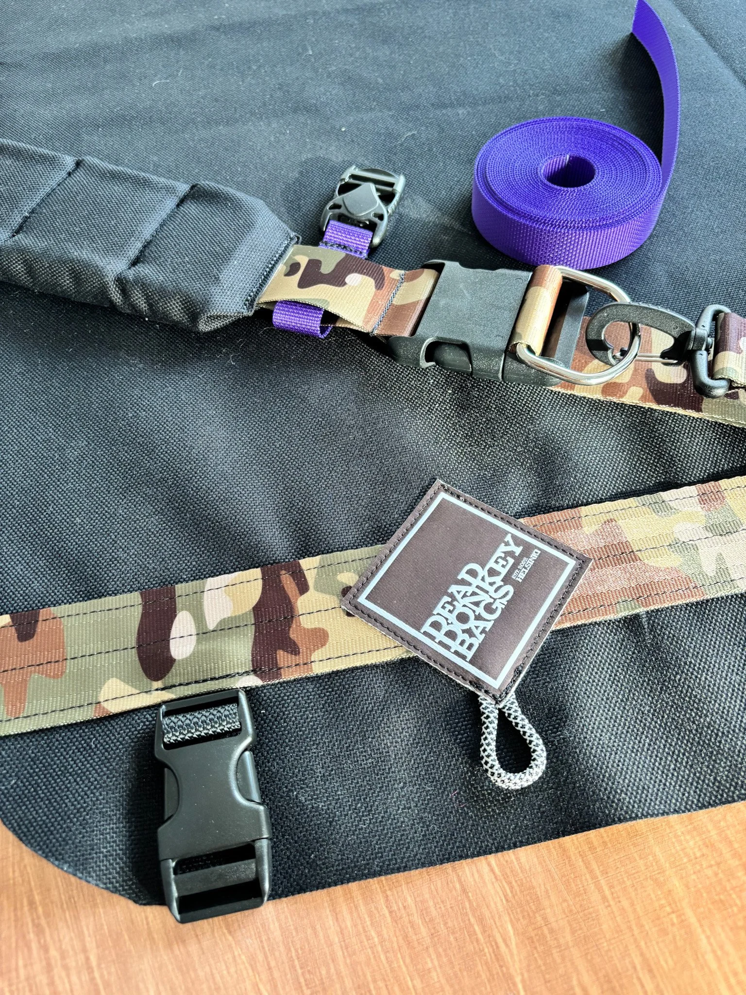 Close-up of a black and tan tactical bag with a camouflage strap, purple strap, and a purple roll of strap, on a wooden surface.