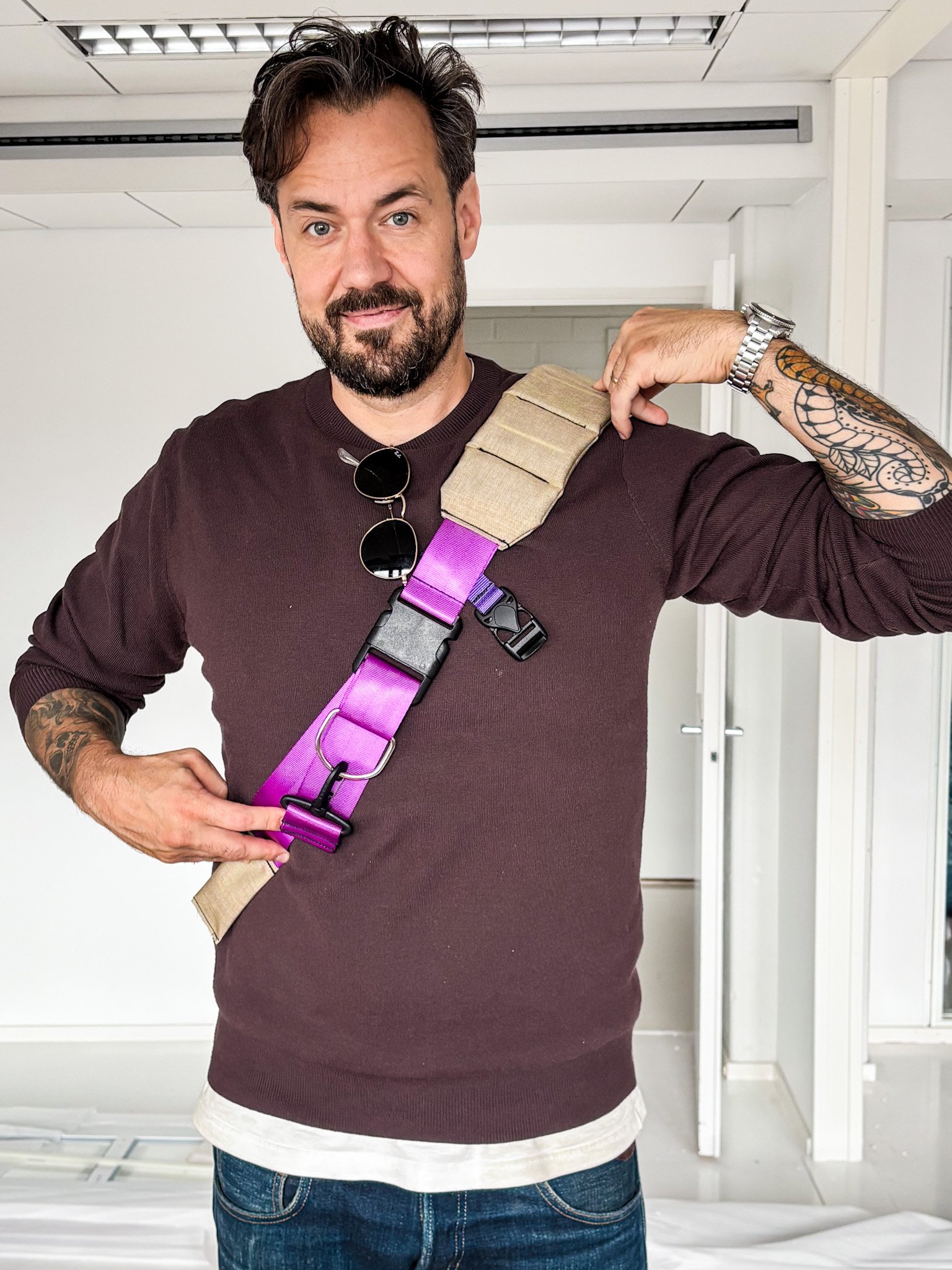 A man with dark hair and a beard is smiling and holding a colorful harness over his shoulder. He is wearing a maroon sweater, sunglasses hanging from his shirt collar, a silver watch, and has tattoos on his arms. He is standing indoors with white walls and a door in the background.