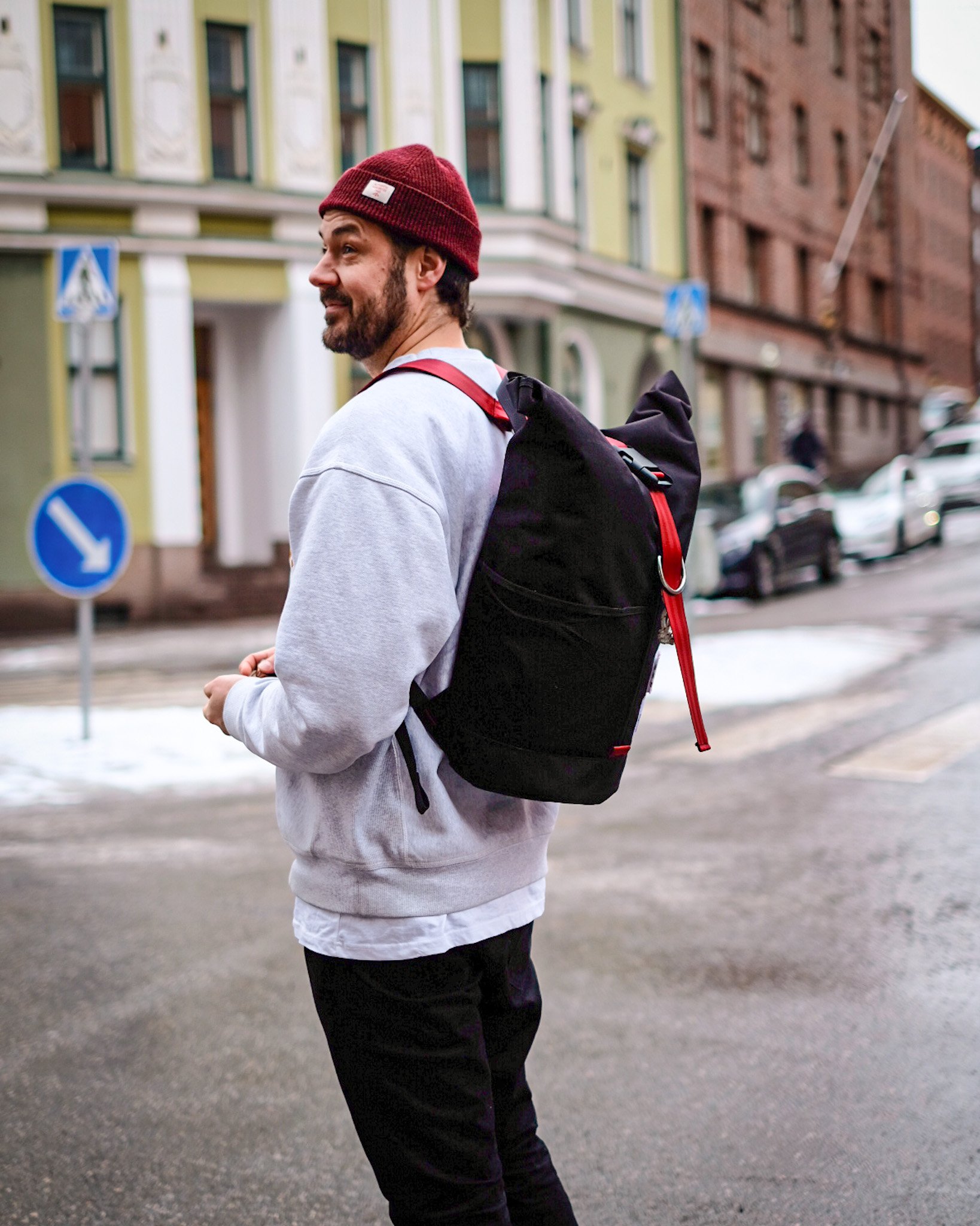 Man with beard wearing red beanie, gray sweatshirt, and black backpack standing on a city street with colorful buildings and parked cars.