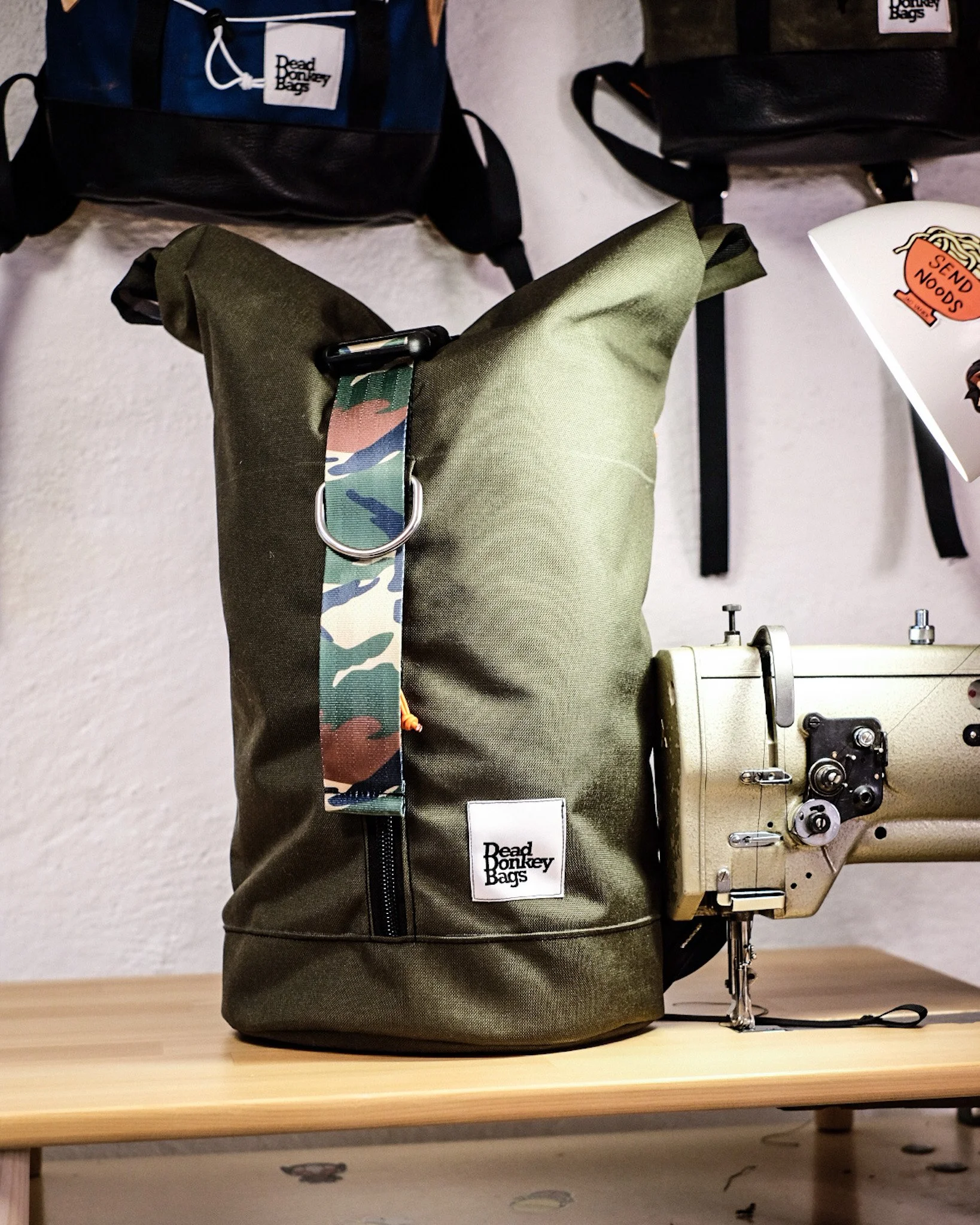 A green backpack with a Dead Donkey Bags label, sitting on a wooden table, next to a vintage sewing machine. There are backpacks hanging on the wall behind.