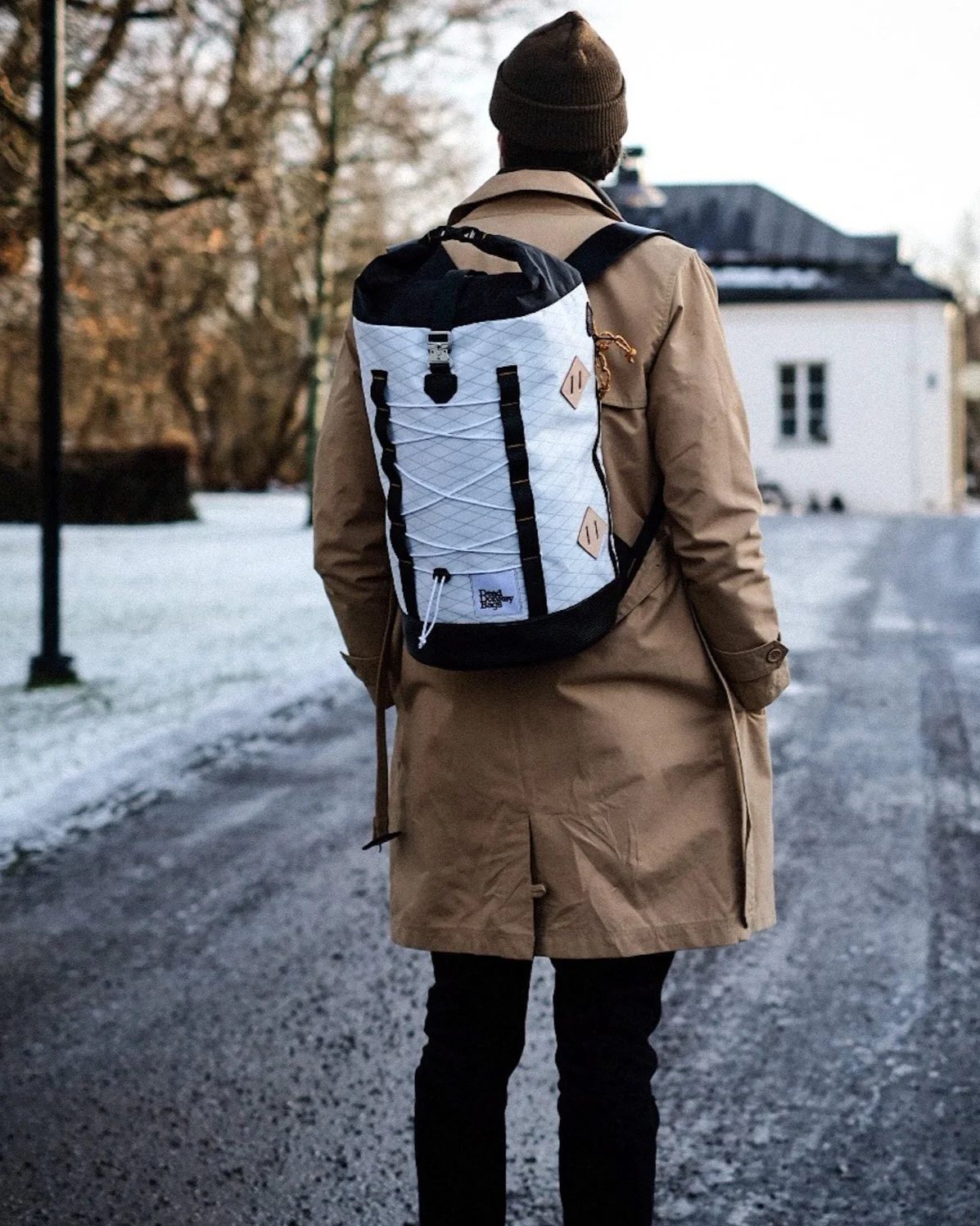 A person wearing a brown coat and a beanie hat, carrying a white and black backpack, walking on a snowy road with trees and a white building in the background.