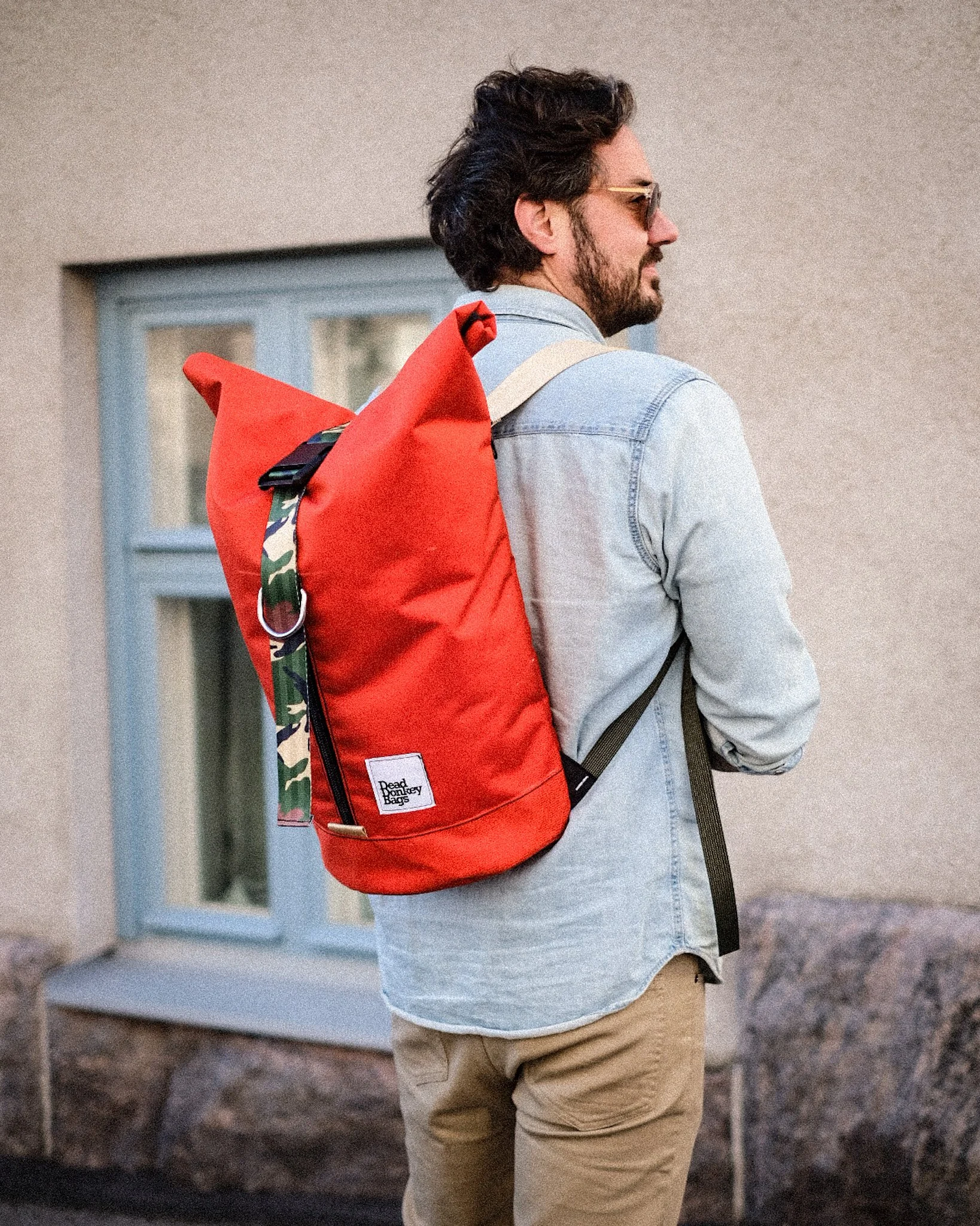A man with dark hair and a beard wearing sunglasses, a light denim jacket, and beige pants, carrying a large red backpack with camouflage accents on the straps, standing outdoors near a house with a blue window.