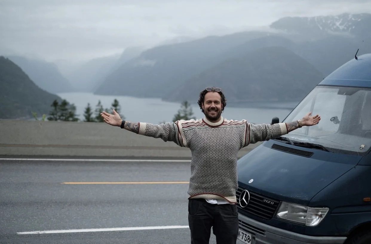 A man standing on a highway with arms outstretched, smiling, next to a black Mercedes-Benz van, with mountains and a lake in the background.
