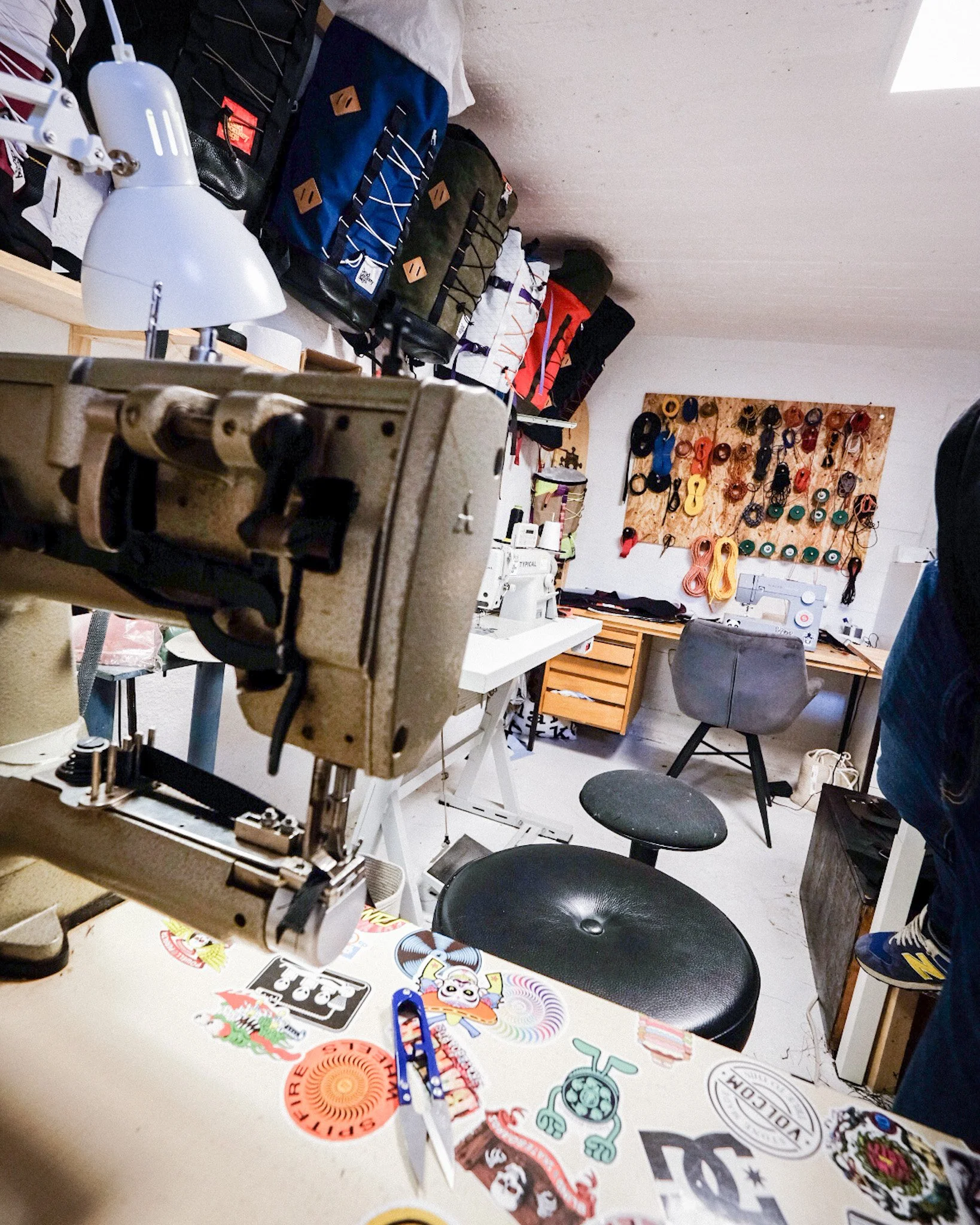 A workshop with a sewing machine on a table, colorful patches and patches on the table, backpacks hanging on the wall, and a pegboard with sewing supplies and tools in the background.
