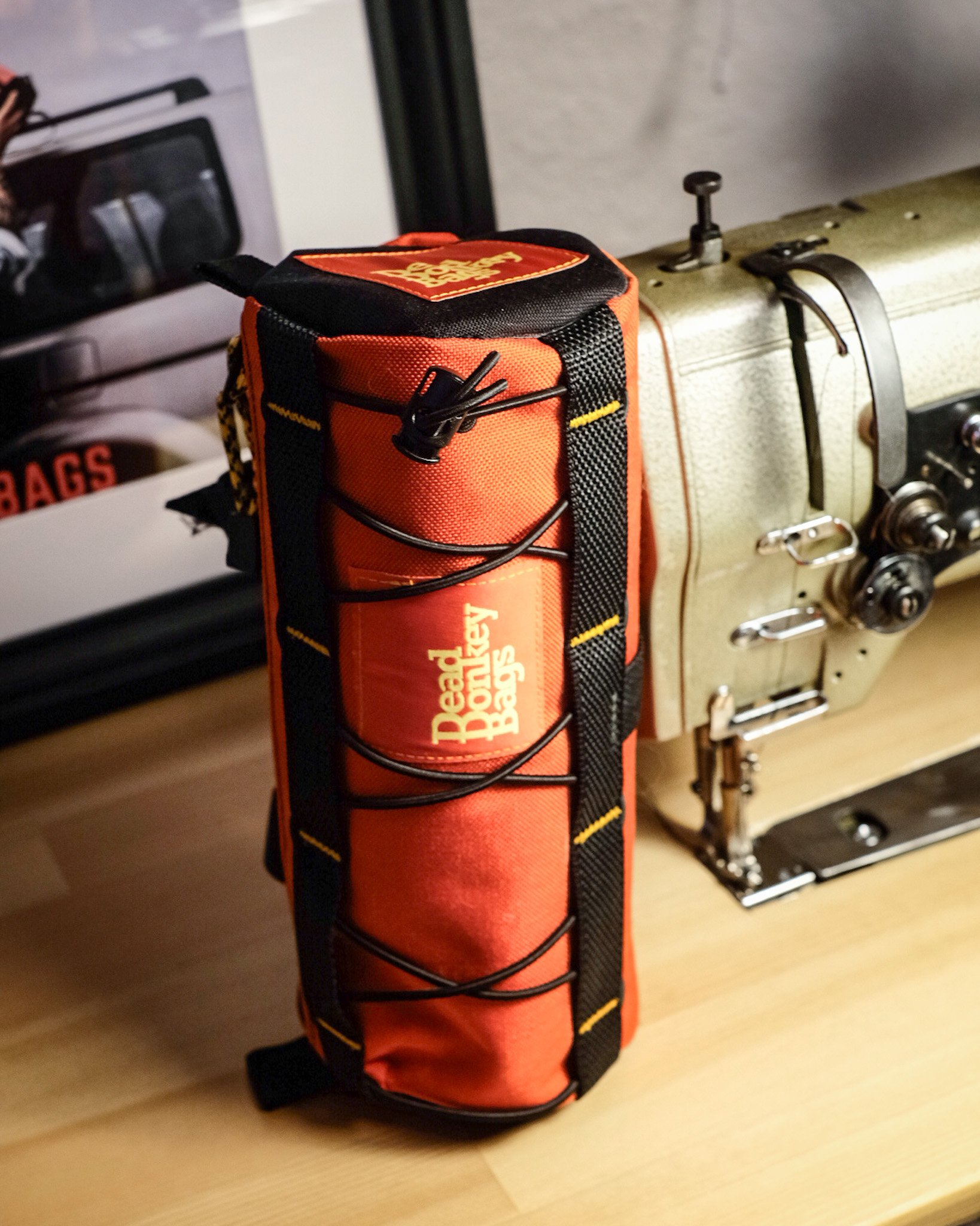 A red and black Leave No Trace Emergency Bag with bungee cord on a wooden table next to a vintage sewing machine.