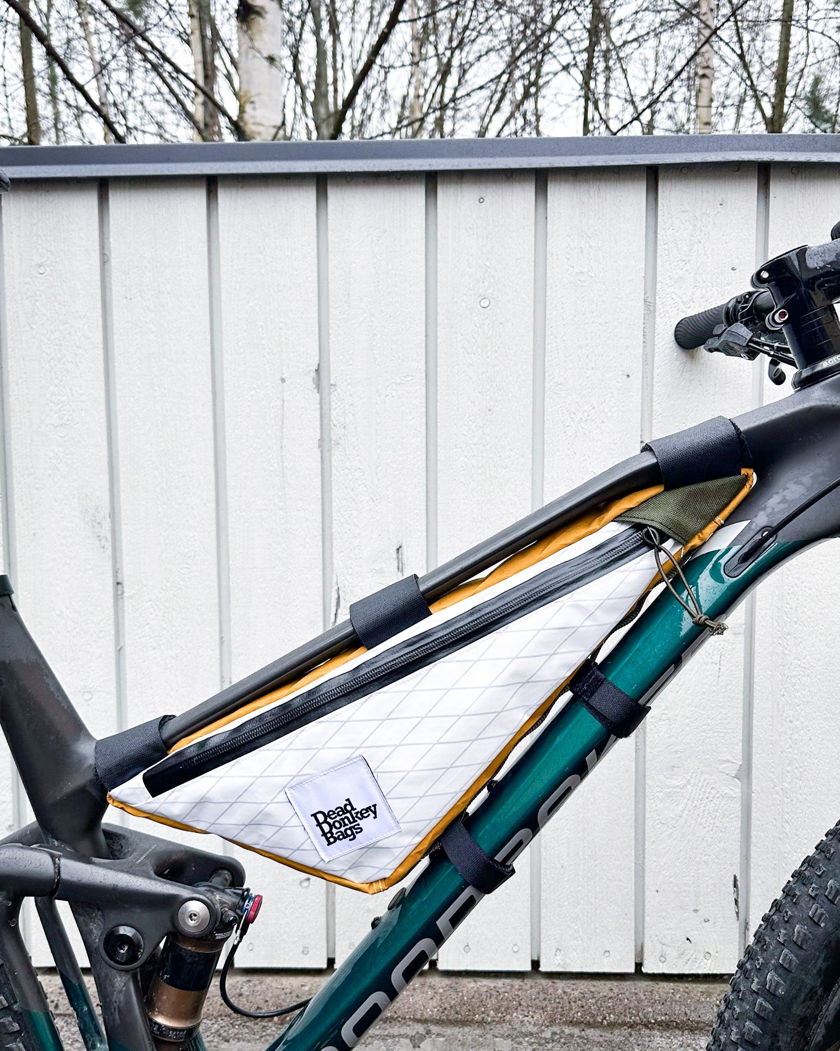 A mountain bike with a white bag attached to its top tube, mounted with black straps, against a white wooden fence background.