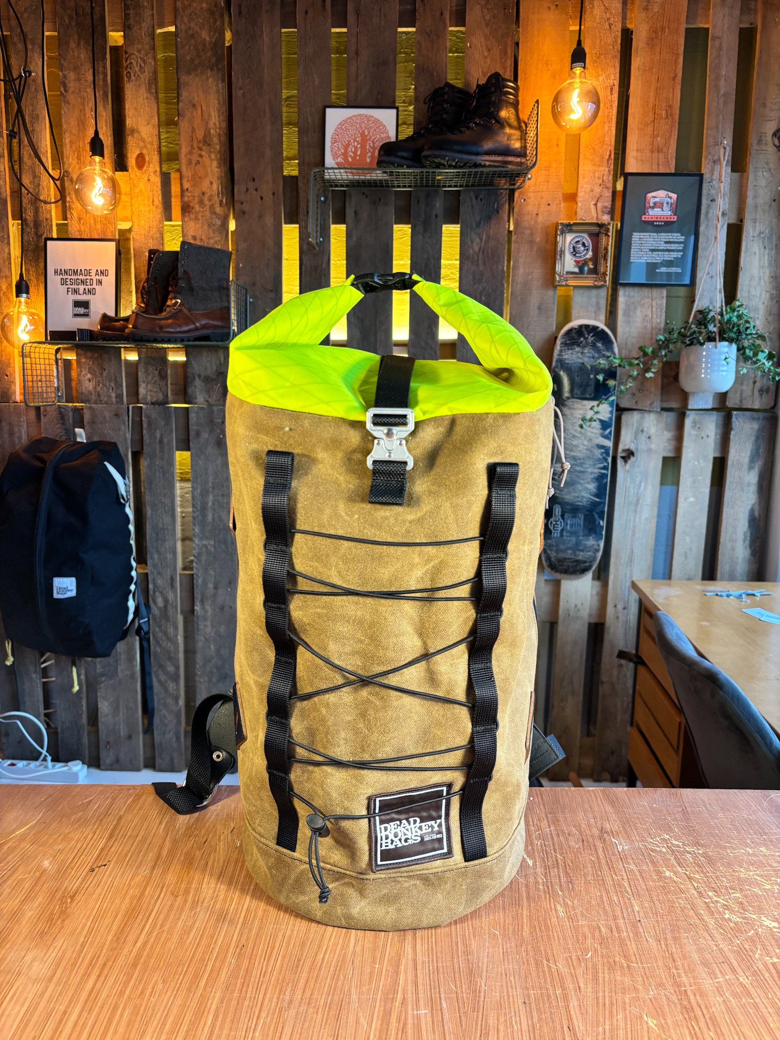 Beardy Bags yellow and brown backpack with black straps and bungee cord on front, with green fabric handle on top, placed on a wooden table.