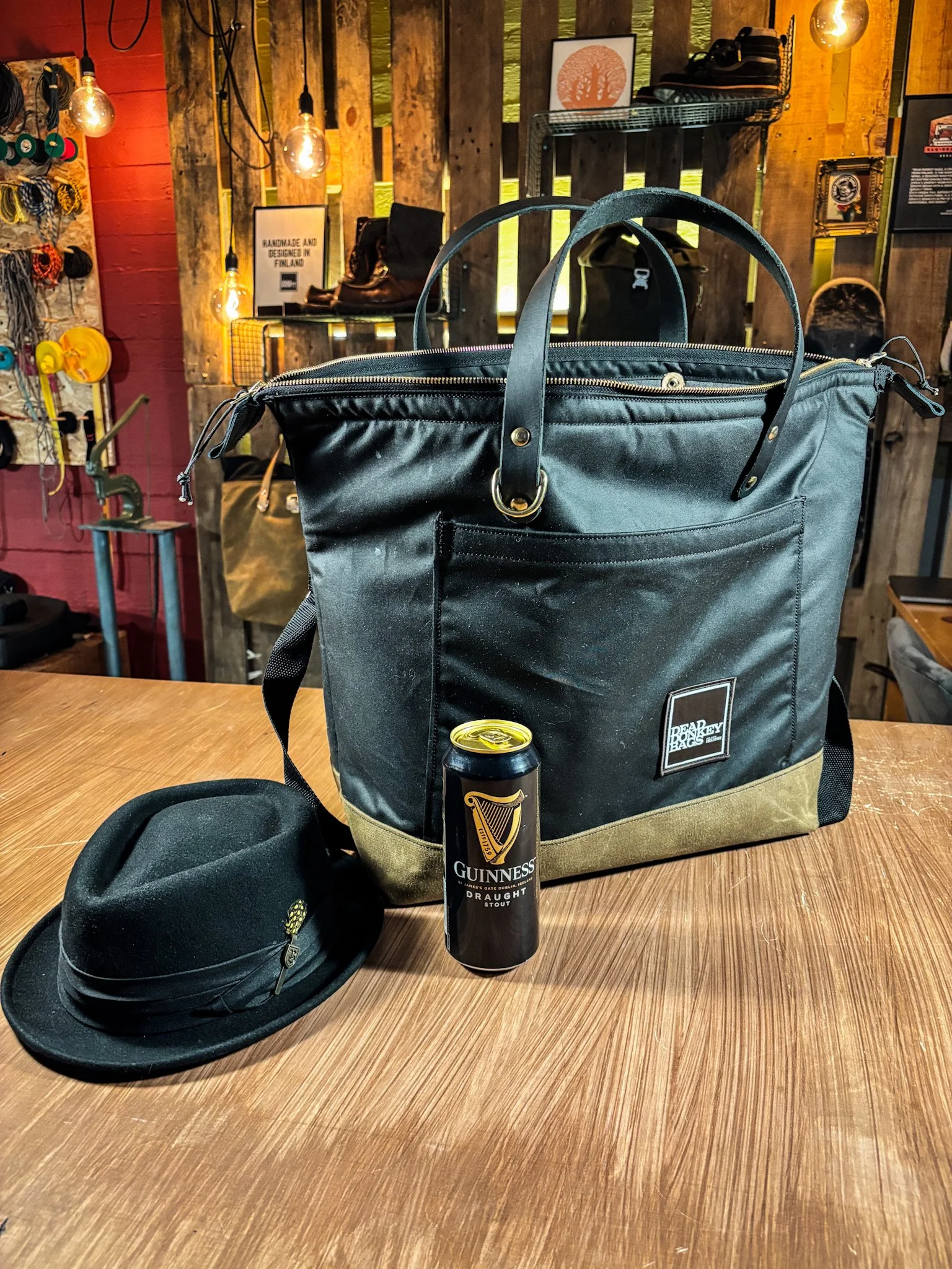 A black Bear Backpack, a black Guinness Draught can, and a black hat with a golden pin are placed on a wooden table. The background features a rustic interior with wooden walls, hanging lightbulbs, and various decorative items.