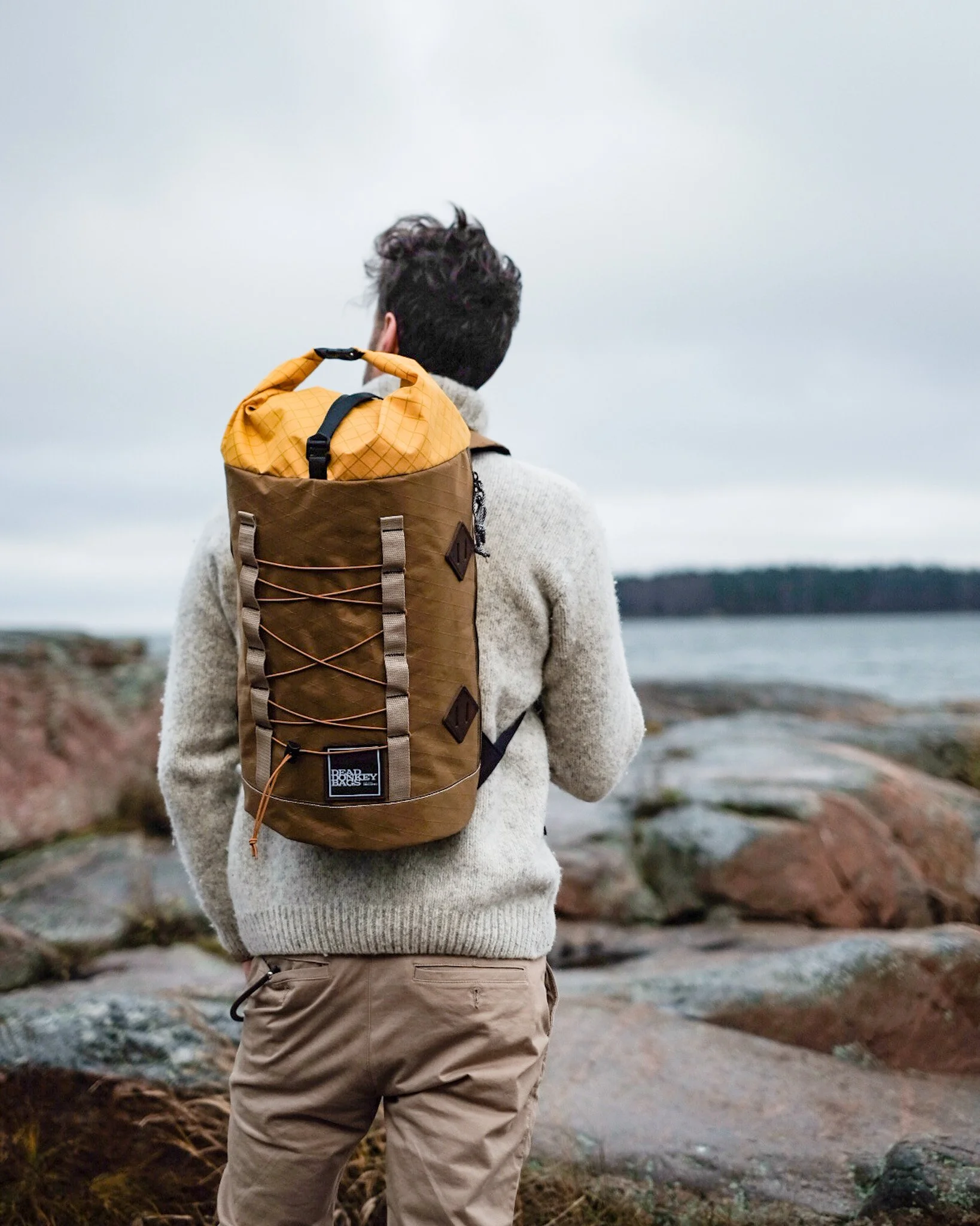 A person standing on rocks near a body of water, facing away, wearing a grey sweater and beige pants, carrying a brown and yellow backpack.