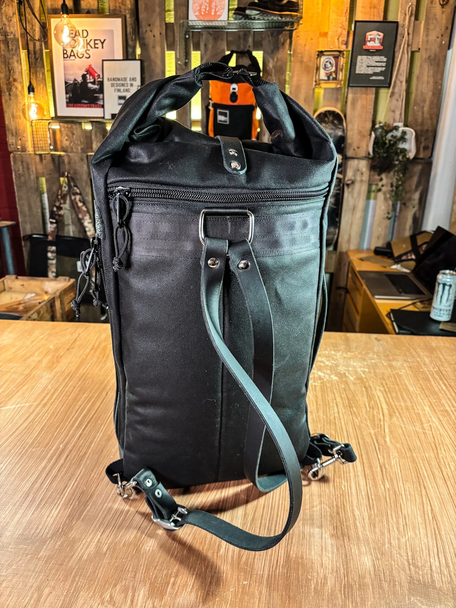 Black backpack with multiple compartments and straps, on a wooden table in a rustic store or cafe setting.