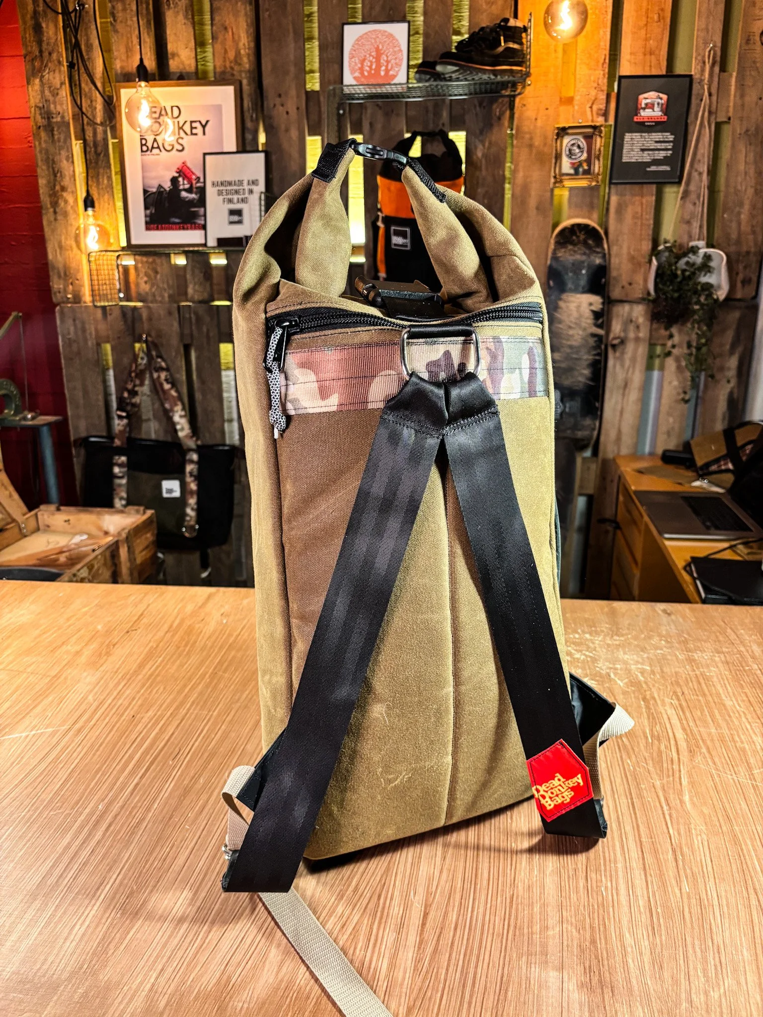 Backpack with padded shoulder straps and various pockets, placed on a wooden table in a store or workshop with wooden walls, framed posters, and other outdoor gear.