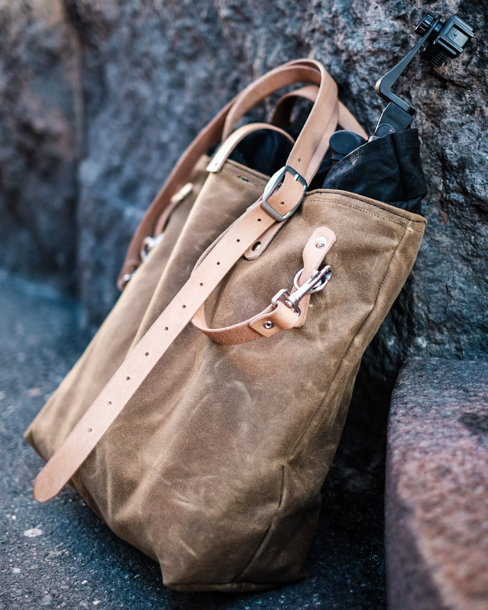 Two tan canvas and leather tote bags resting against a rough stone wall, with one bag holding a black umbrella with a curved handle.