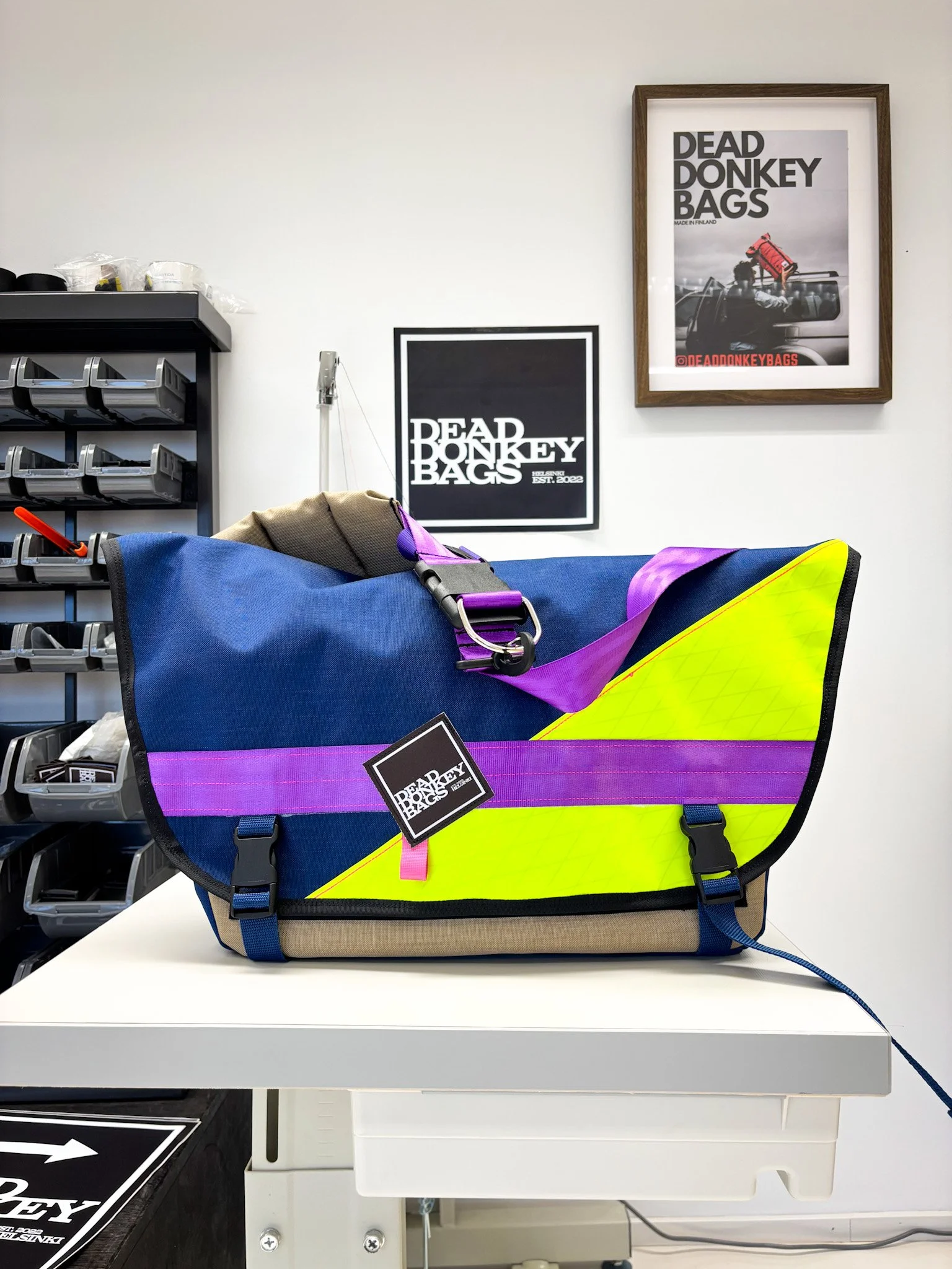 Colorful duffel bag with purple, yellow, and blue sections on a white table in a workshop or store. Two framed posters of Dead Donkey Bags on the wall behind it, with storage bins on a shelf to the left.