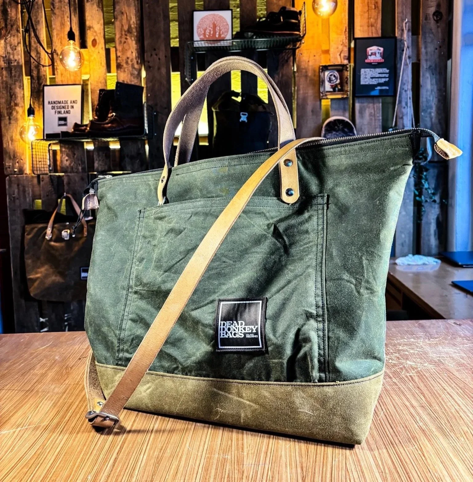 A green canvas tote bag with leather handles and a shoulder strap sitting on a wooden surface inside a rustic store with wooden walls and shelves displaying various bags and accessories.