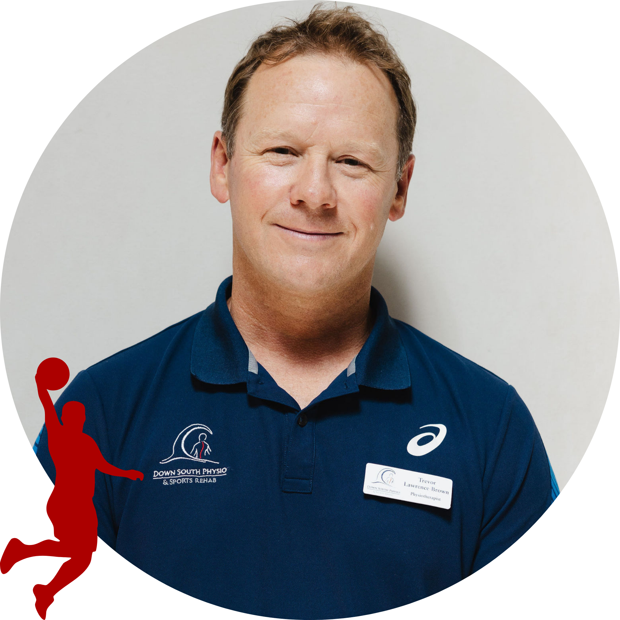 A man with short, light brown hair, wearing a navy polo shirt with a logo for Down South Physio & Sports Rehab and a name tag that reads Trevor Lawrence Brown, standing against a light background and smiling.