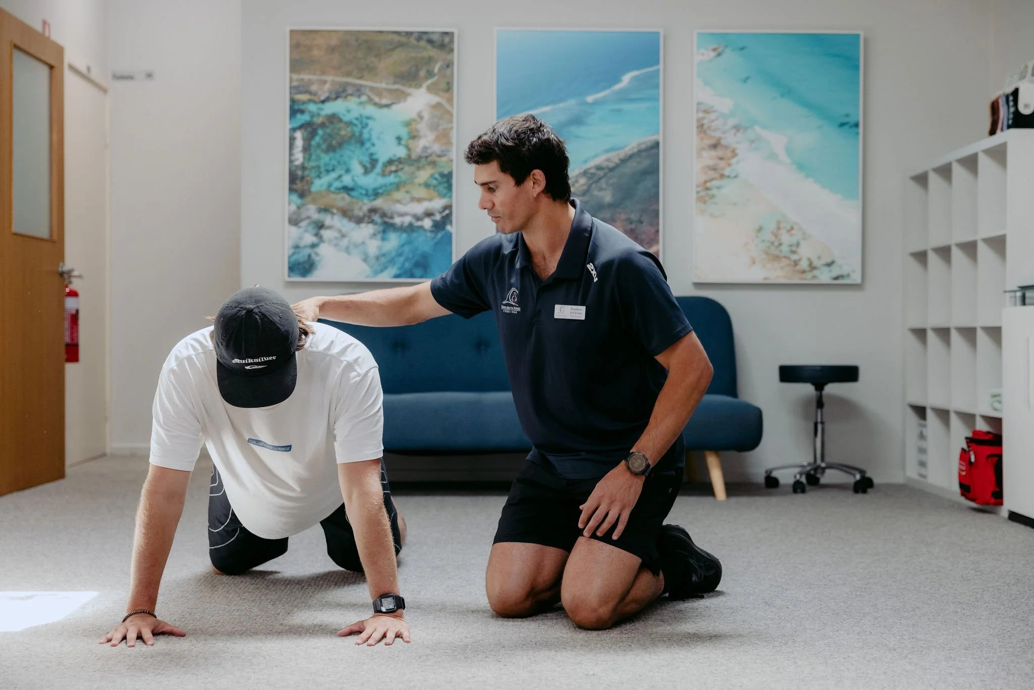A man in a white T-shirt and black shorts performs a push-up on the carpeted floor while kneeling on one knee. A person in a navy shirt and black shorts gently places a hand on the other person's head, providing support or assistance. The room features a navy couch, three large landscape wall art pieces, a white shelf, and a stool.