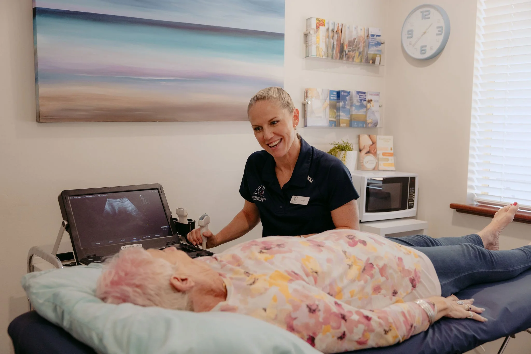 A healthcare professional performs an ultrasound on an elderly woman lying on an examination table in a medical office.