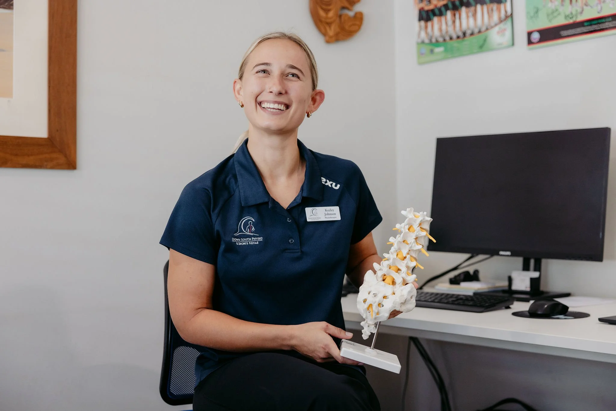 A woman wearing a navy polo shirt with a name tag, sitting in an office, holding a model of the human spine with a smile.
