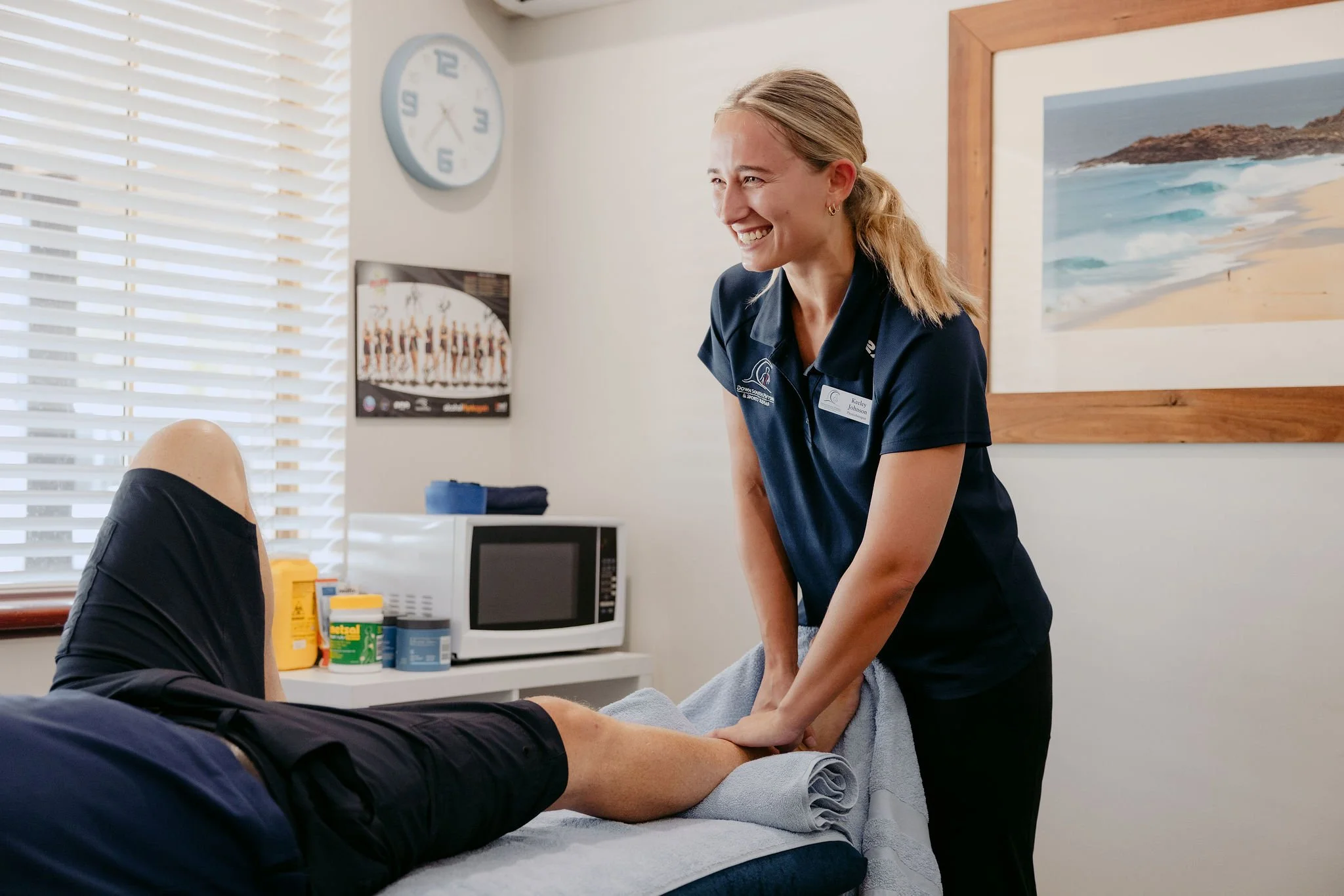 A woman giving a massage to a person lying on a massage table in a treatment room. The woman is smiling and wearing a navy blue uniform with a name tag. The room has a window with blinds, a wall clock, a framed beach picture, and medical supplies on a side table.
