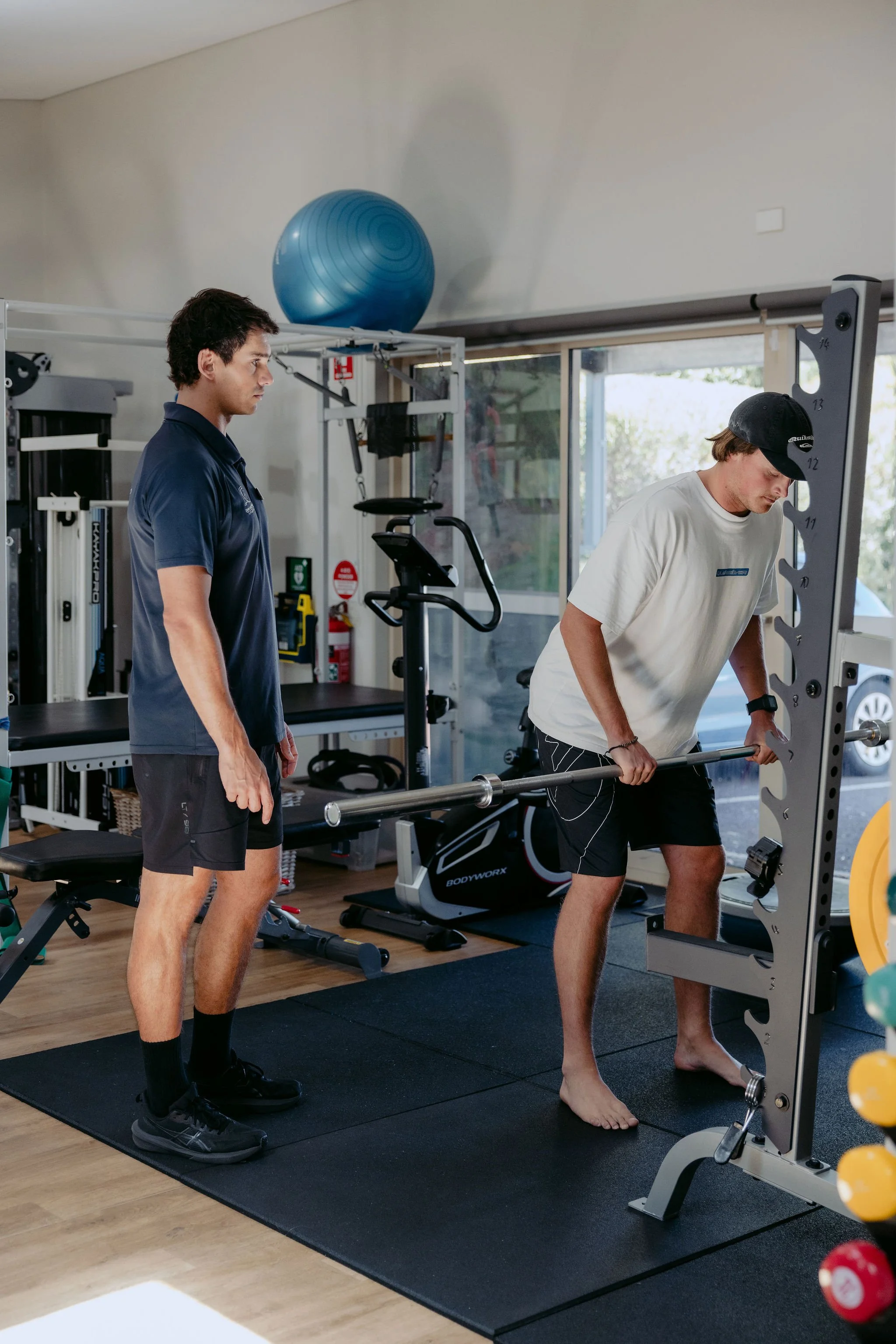 A young man performing a barbell exercise under the supervision of a trainer in a gym.