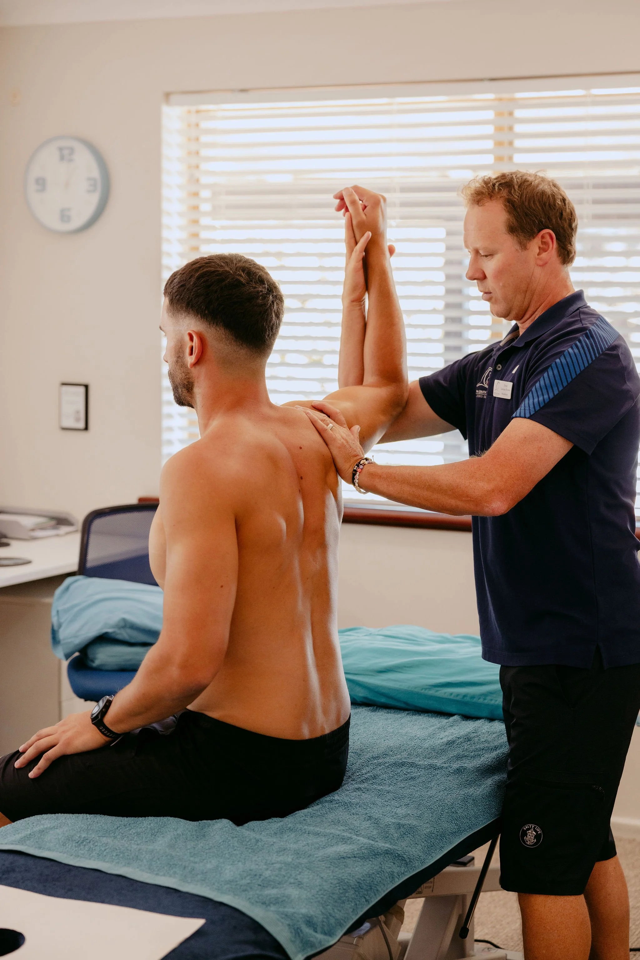 A physical therapist performing a shoulder stretch on a seated shirtless man in a clinic room.