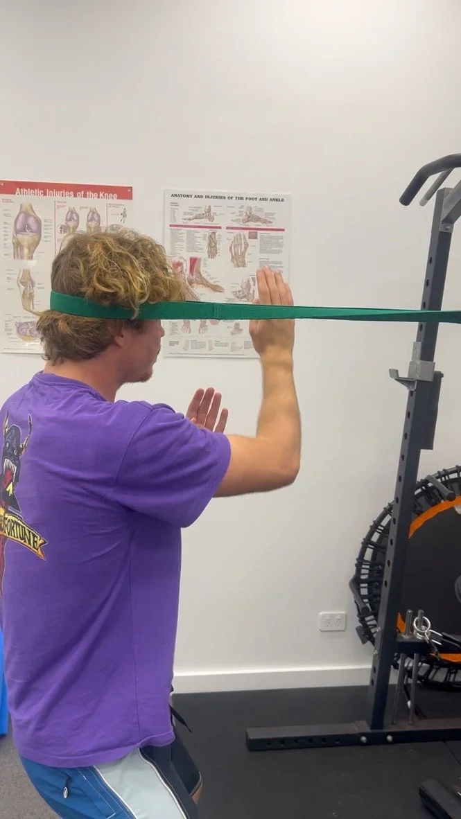 A person with curly hair wearing a purple shirt and shorts is exercising in a gym. They are using a resistance band placed around their head, pulling it while standing next to a workout rack. There are anatomical posters on the wall behind them.