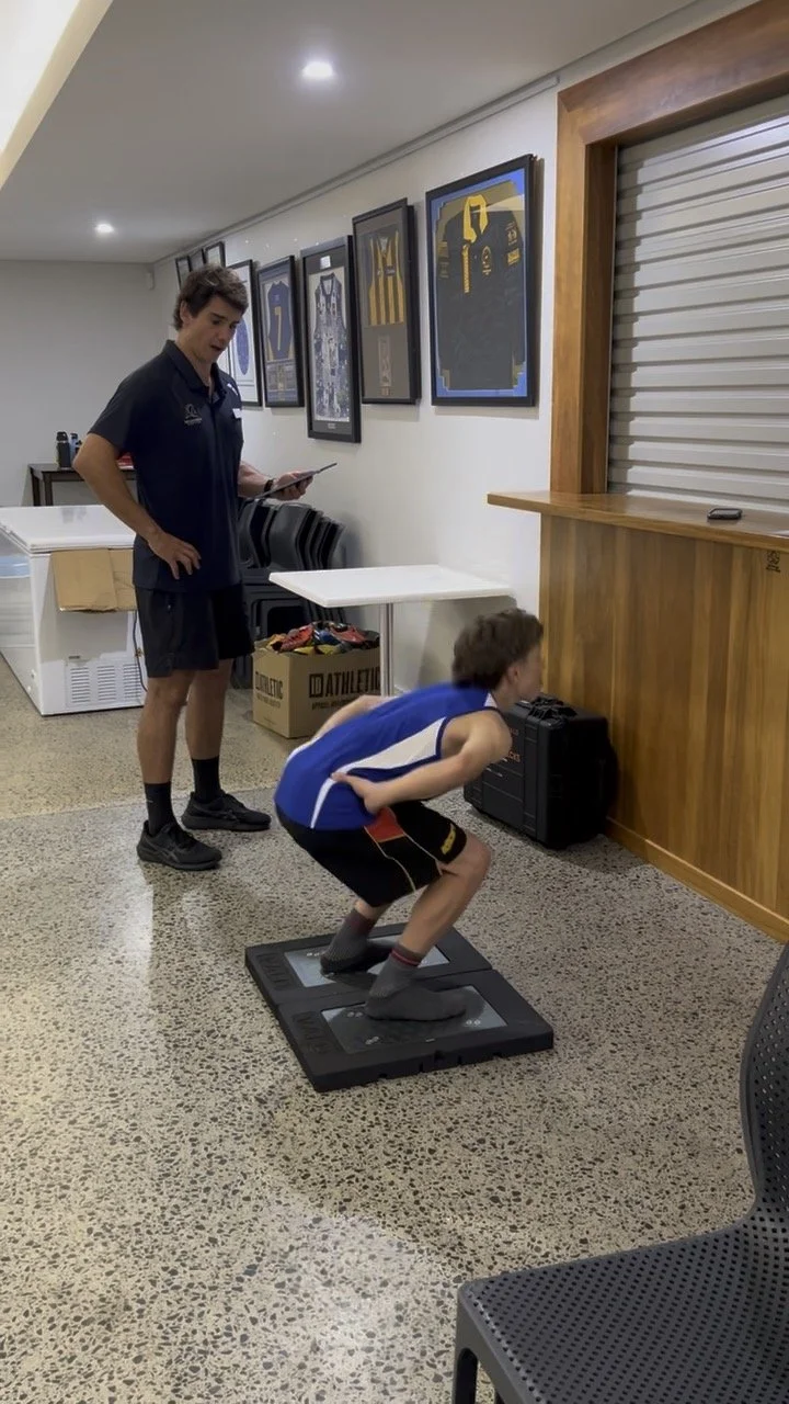 A young boy wearing a blue sports jersey and black shorts squats on a balance board, while a woman in athletic clothing watches and holds a tablet in a room decorated with framed sports jerseys and memorabilia.