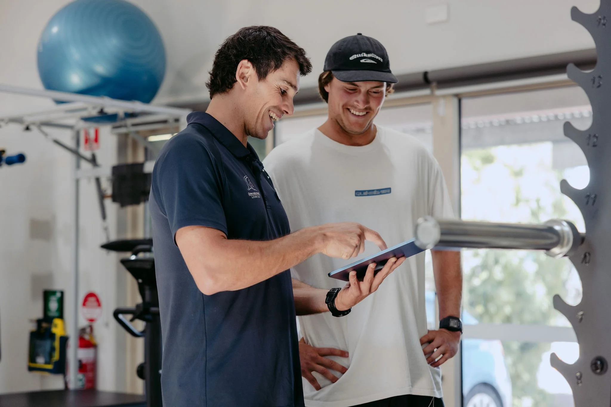 Two men in a gym, one showing something on a tablet to the other, with gym equipment and a large exercise ball in the background.
