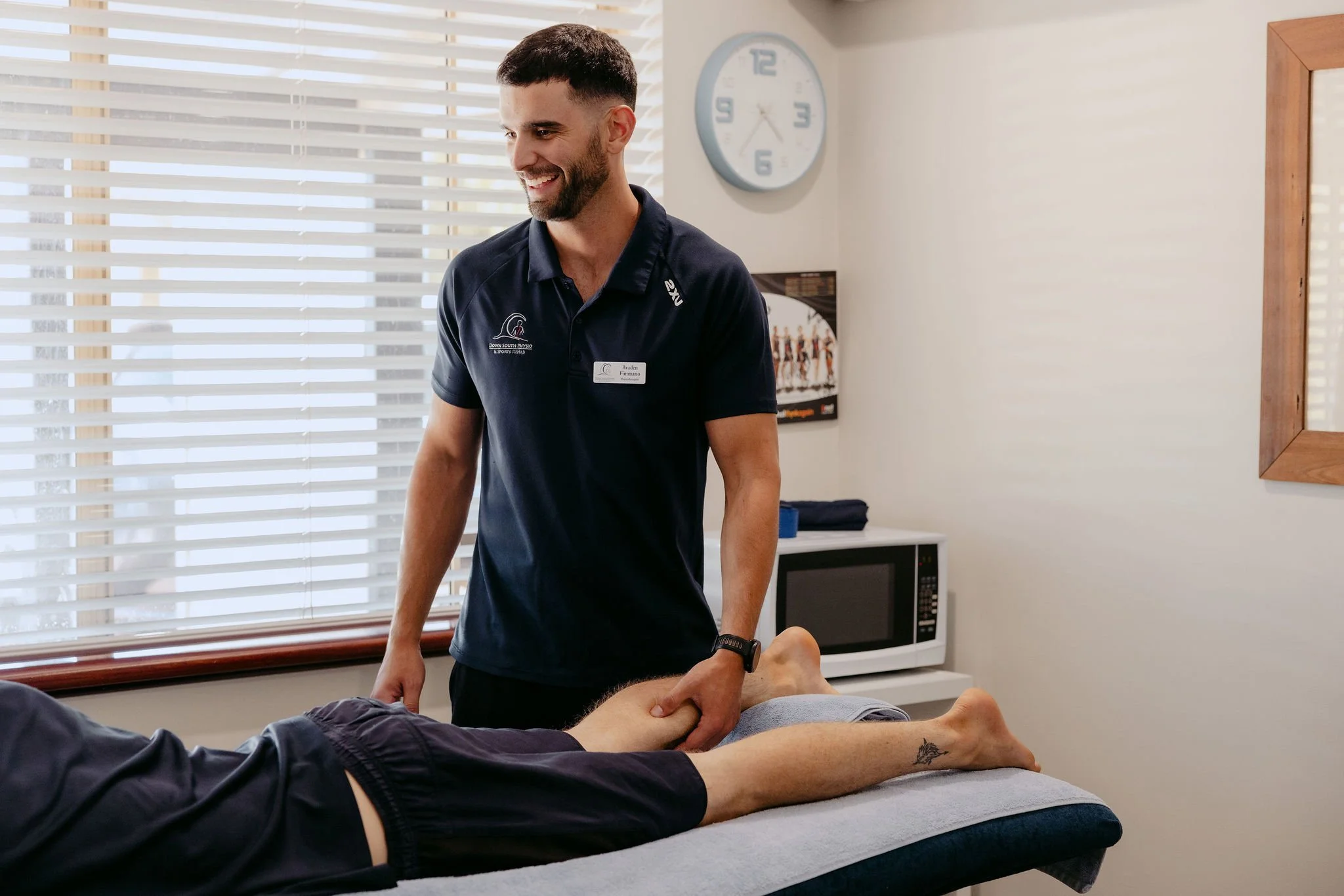 A man receiving physical therapy on his leg from a therapist in a clinic room with blinds, a clock, and a microwave.