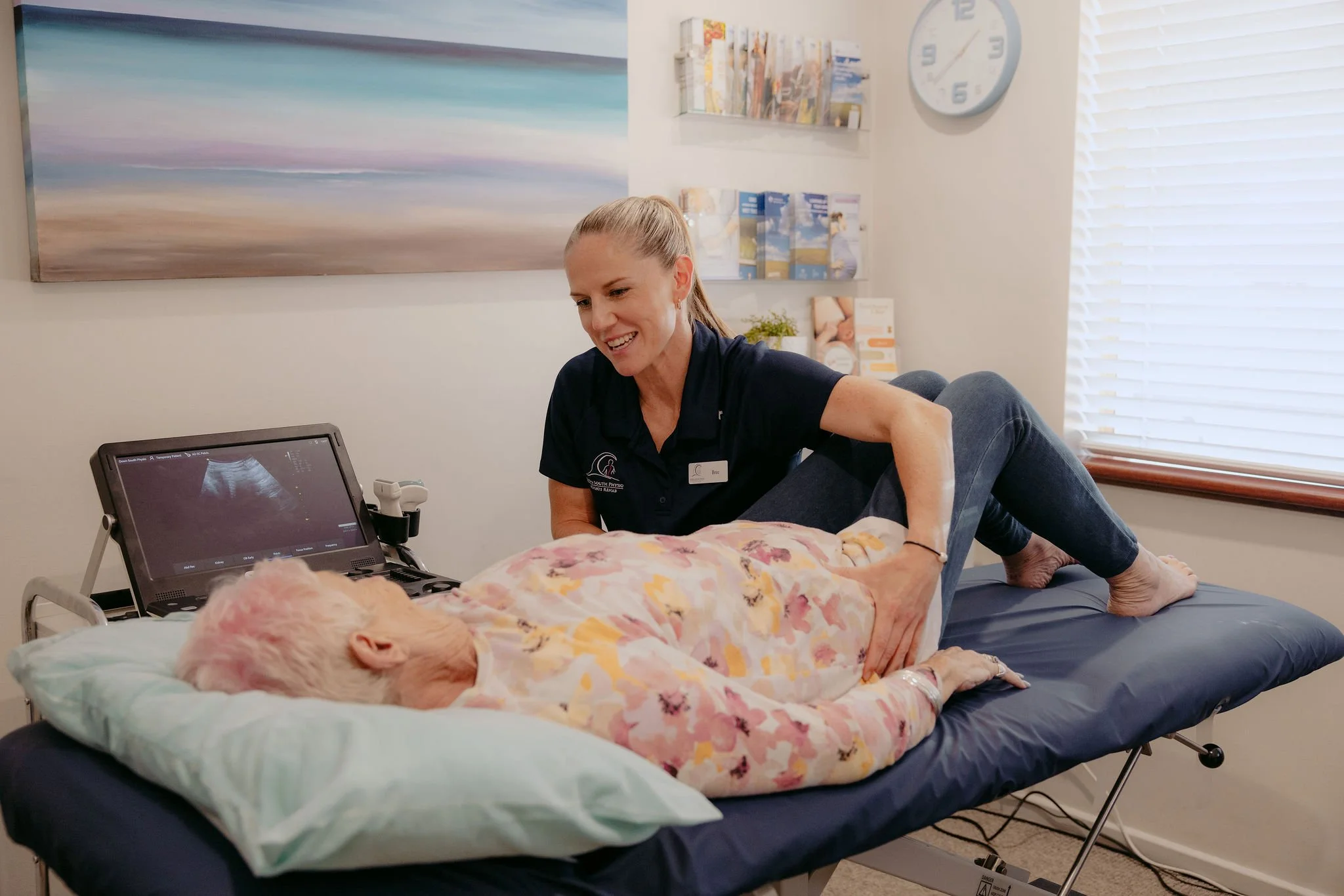 Medical professional performing an ultrasound on an elderly woman lying on an examination table, with ultrasound monitor and medical supplies in the background.