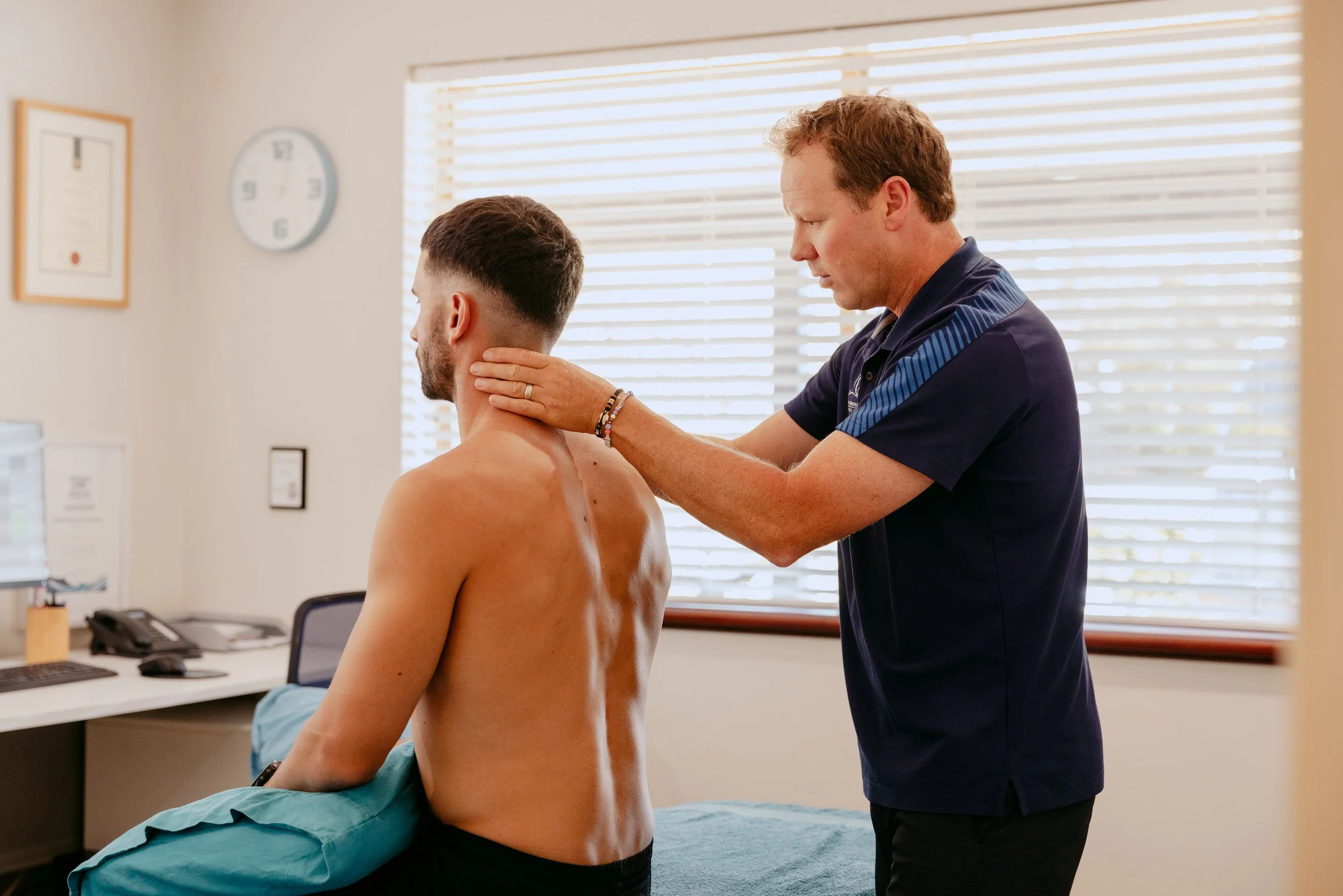 Doctor examining a shirtless man's neck in a clinic room with window blinds.