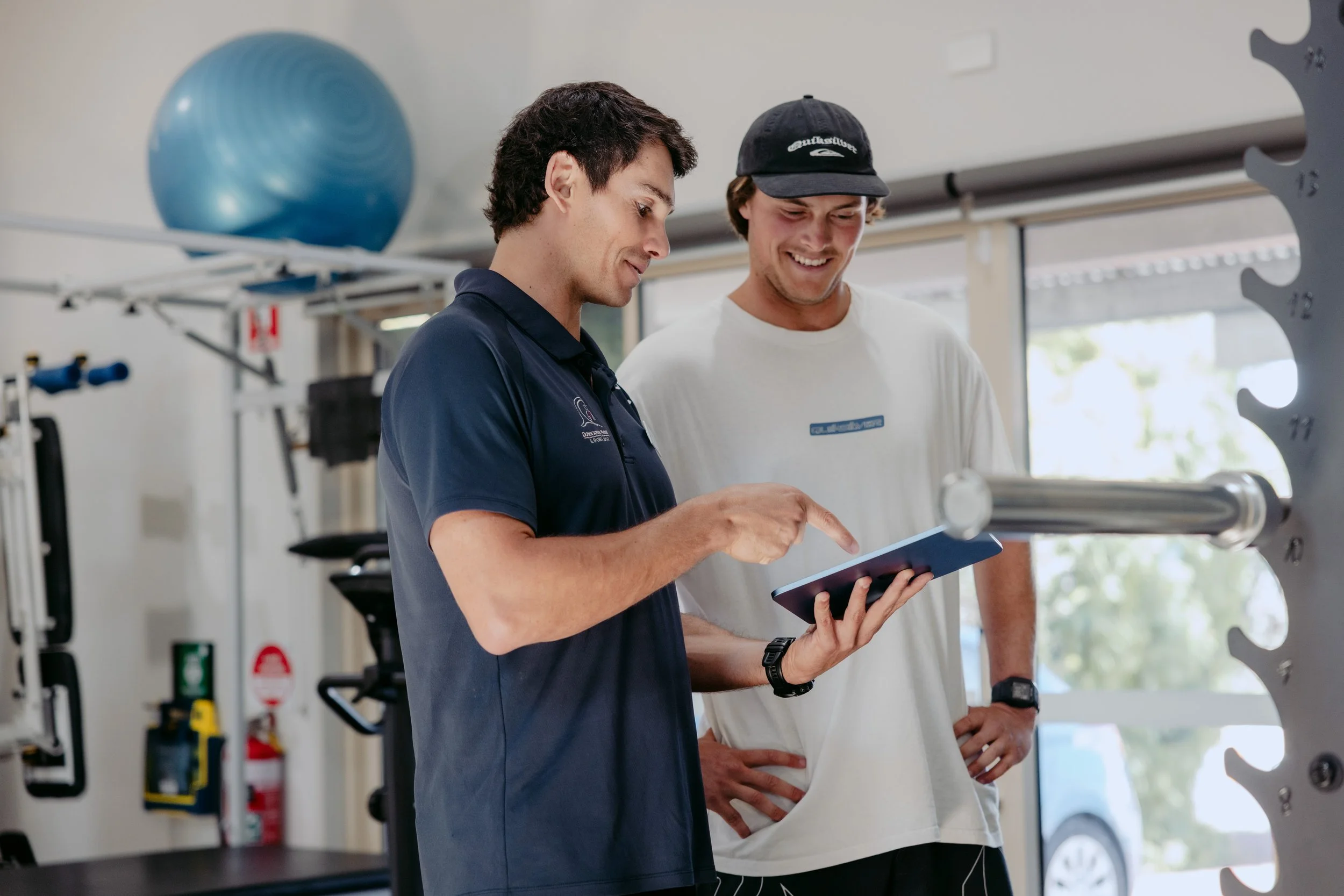 Two men in a gym looking at a tablet, one pointing at the screen, gym equipment in the background.