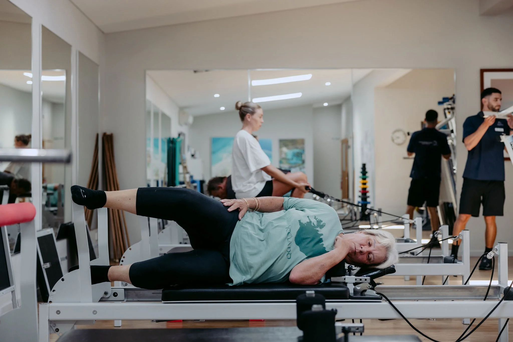 An elderly woman lying on a rehabilitation treadmill, talking on her cellphone inside a physiotherapy clinic with other staff and patients in the background.
