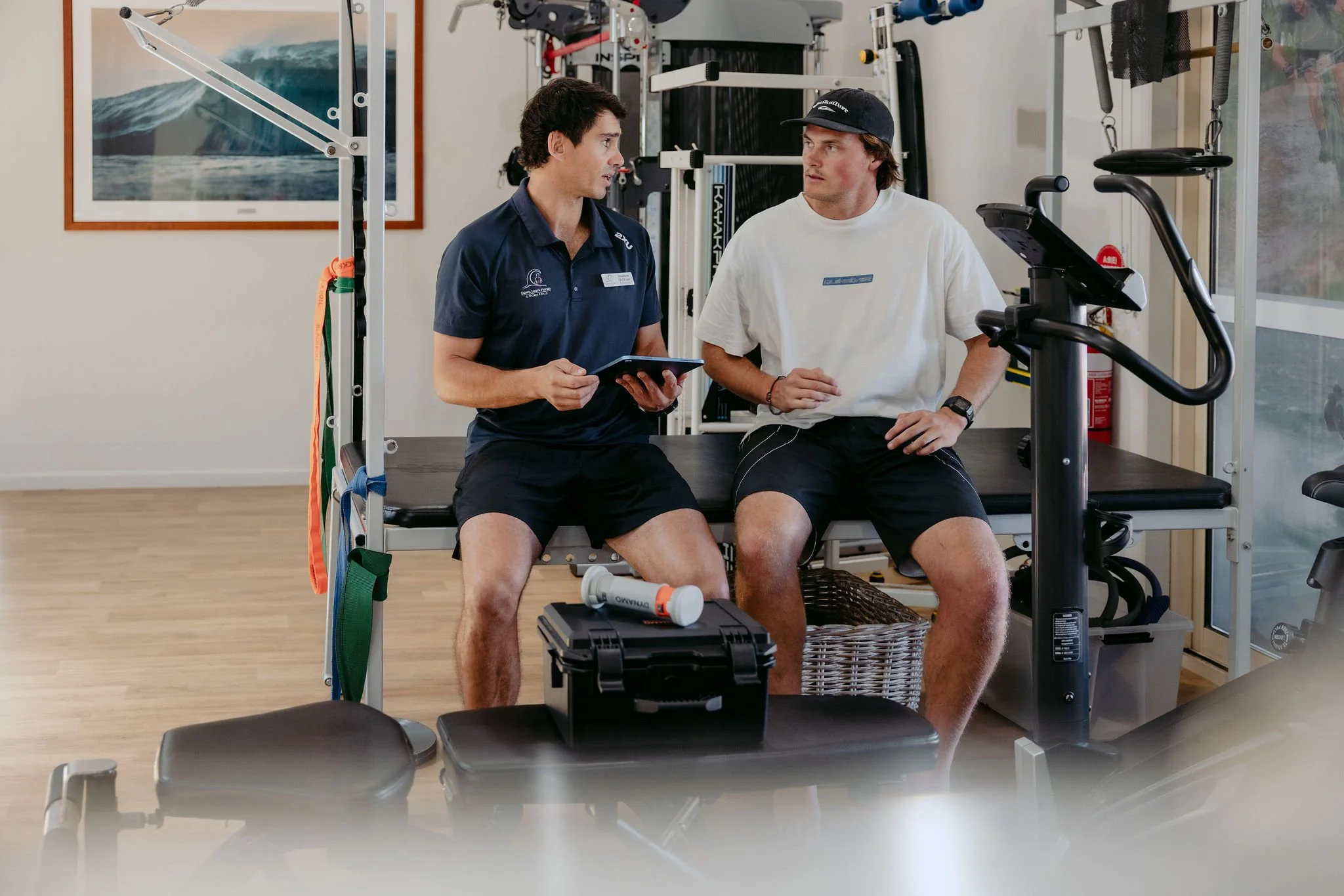 A physical therapist or trainer talking to a male athlete or patient in a gym, sitting on a bench with exercise equipment around them.