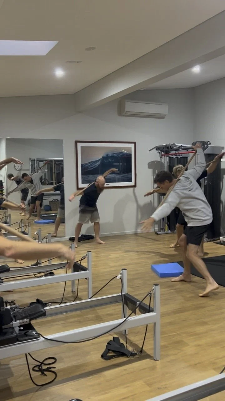 People participating in a Pilates or yoga class using reformer machines, with some holding sticks, in a fitness studio with wooden floors and a painting of a wave on the wall.