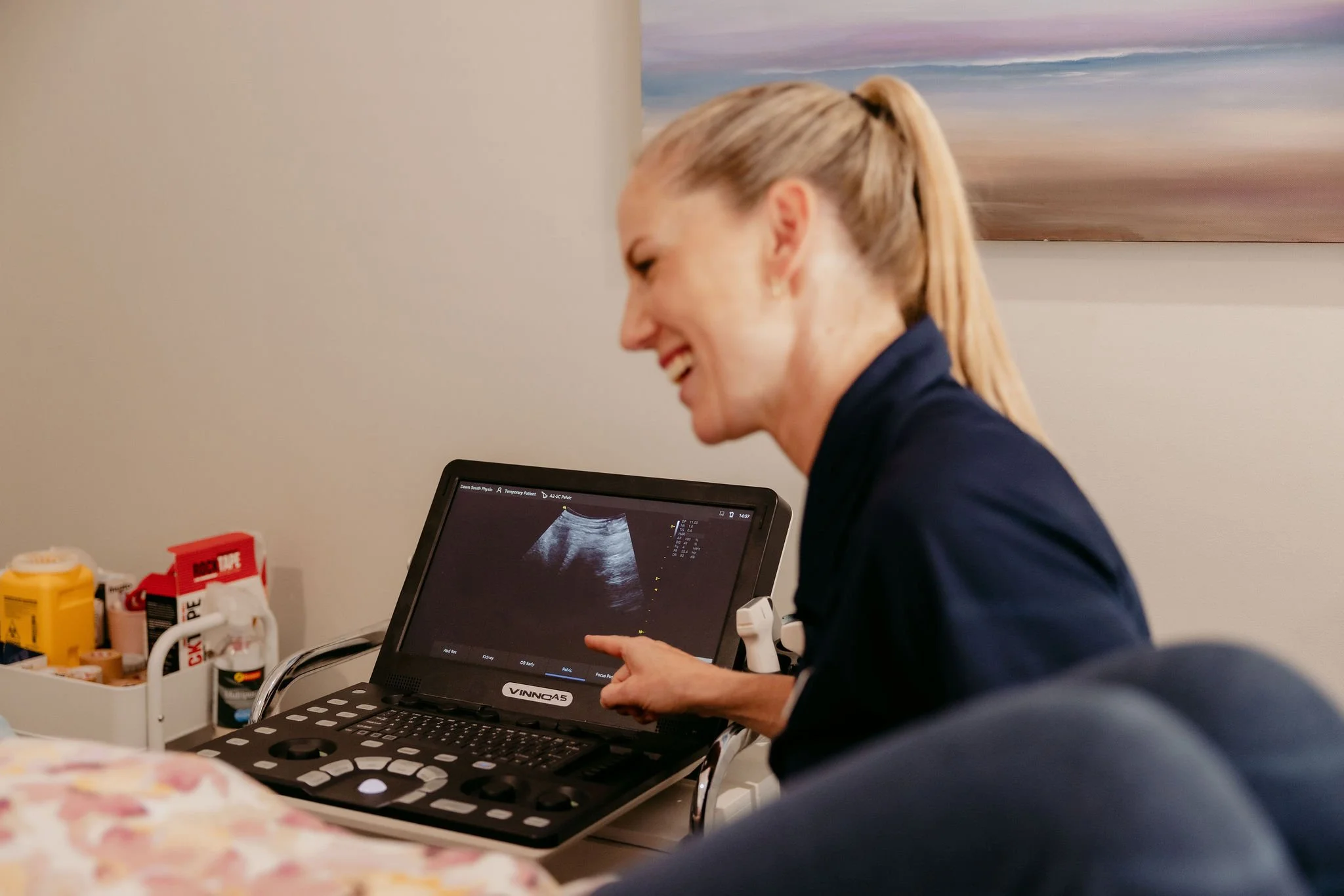 A woman with blonde hair tied in a ponytail looking at an ultrasound monitor and smiling. The ultrasound image is visible on the monitor, and she appears to be in a medical setting with medical supplies in the background.