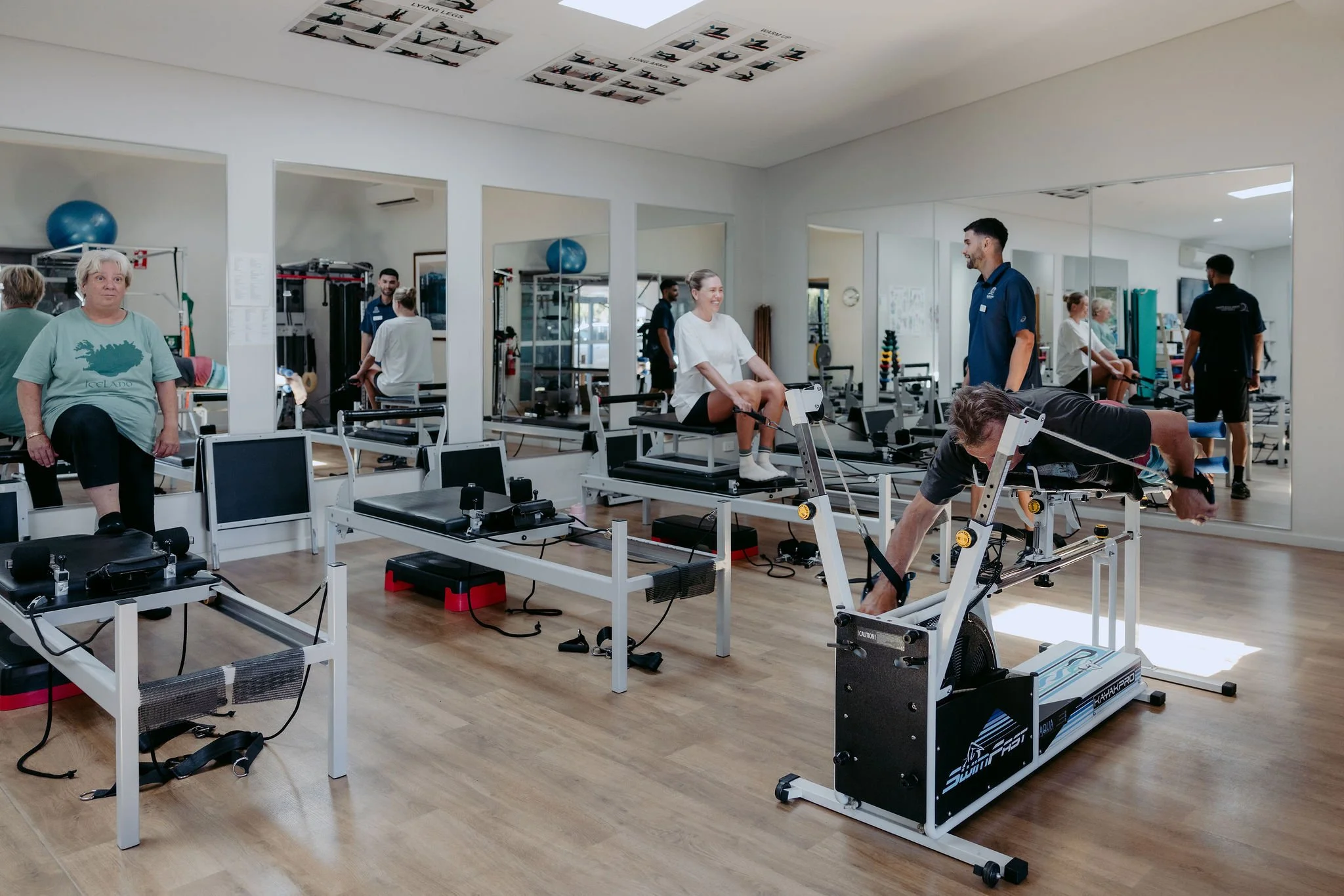 People exercising and stretching in a physical therapy or rehabilitation gym with exercise equipment, mirrors, and a wood floor.