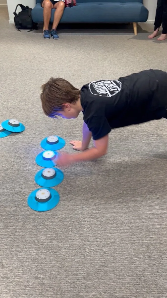 A boy doing push-ups on a carpeted floor with several blue and white devices arranged in a line. There is one person sitting on a blue sofa in the background, and another person's legs are visible on the right side of the image.