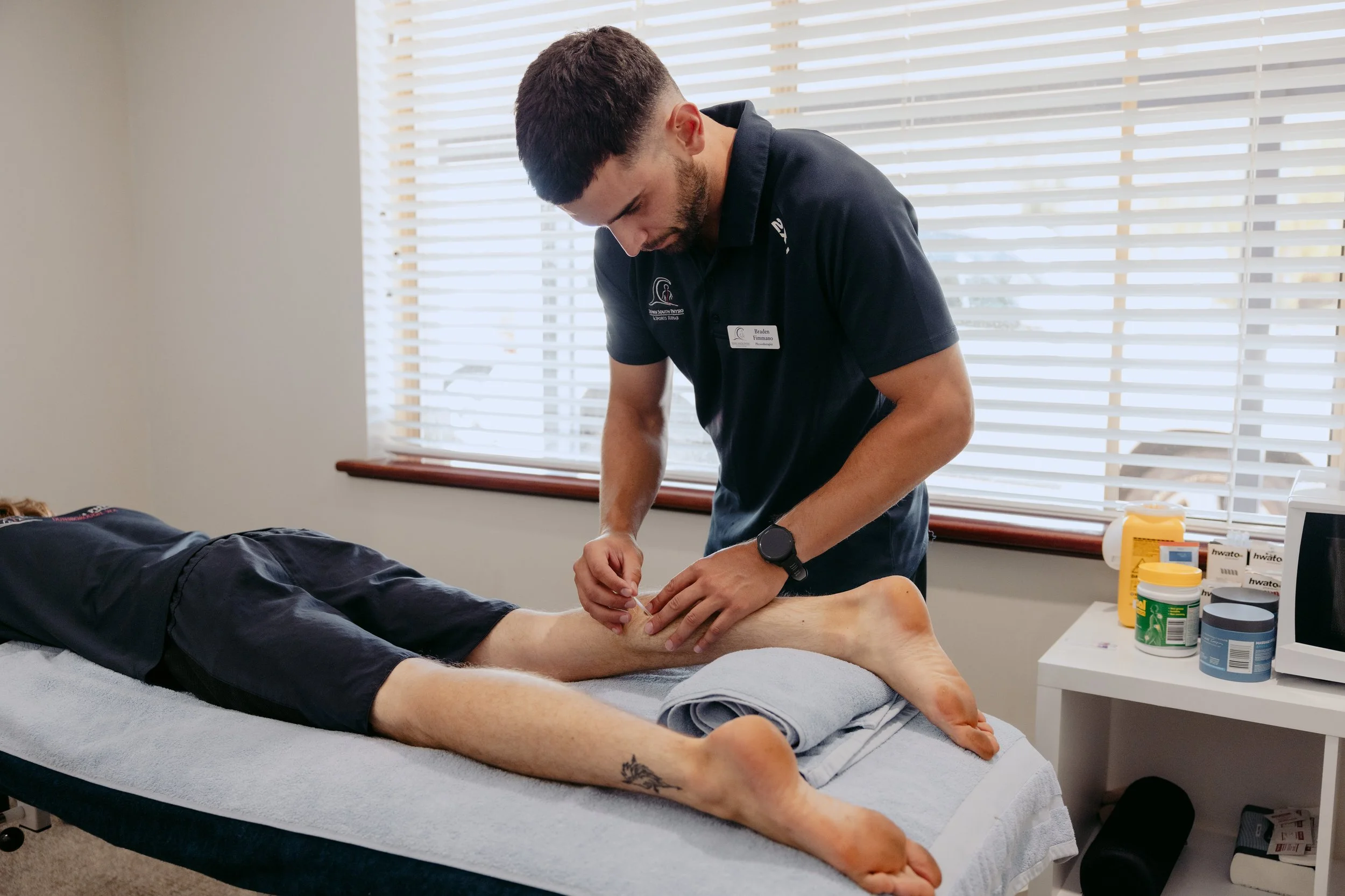 A physical therapist is working on a patient's leg during a therapy session in a well-lit room with window blinds. The patient is lying face down on a treatment table with legs extended, and the therapist is using a needle or similar tool on the lower leg.