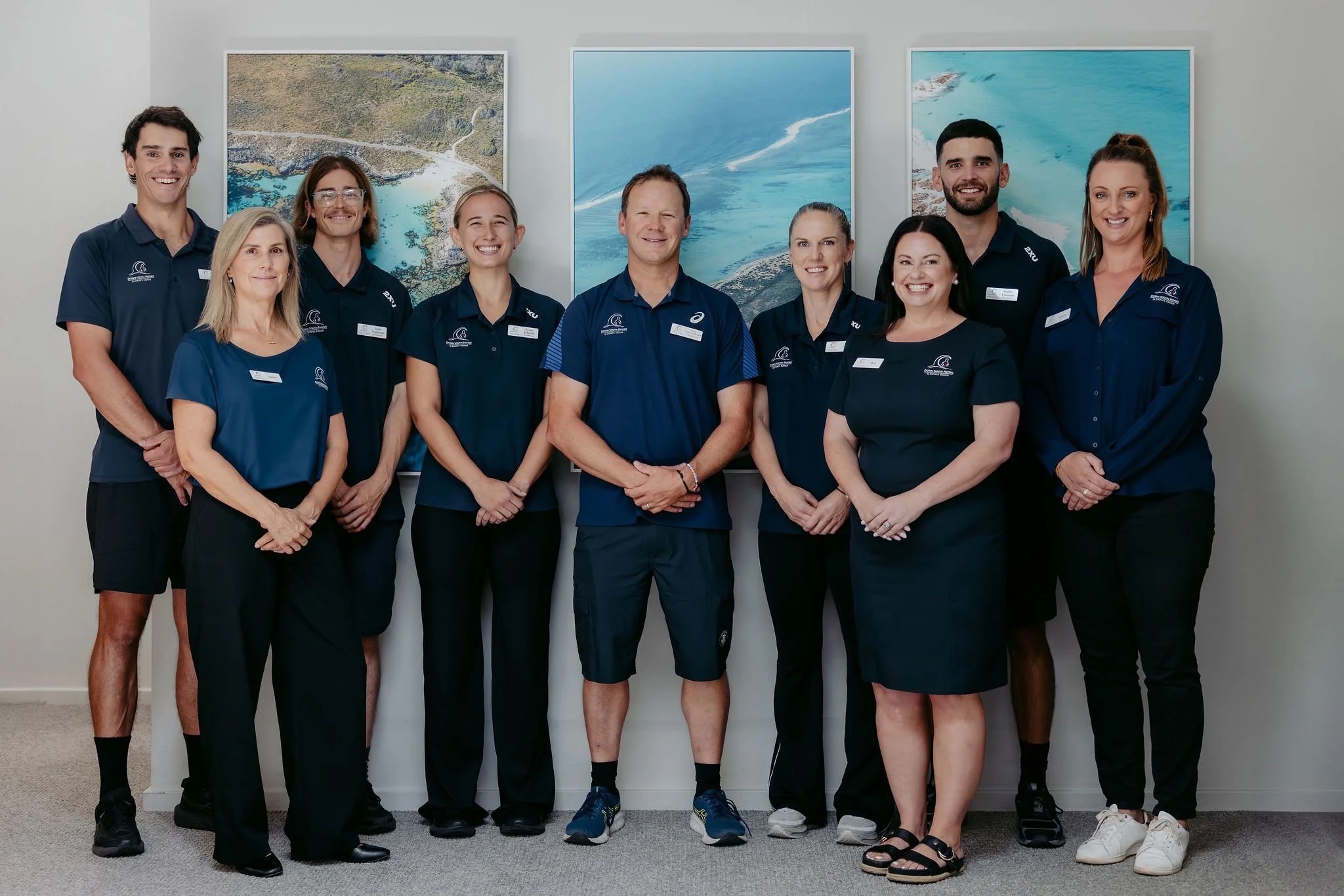 Group of nine people standing in an office, wearing navy blue shirts with wave logos, smiling, with large framed pictures of ocean and coastline on the wall behind them.