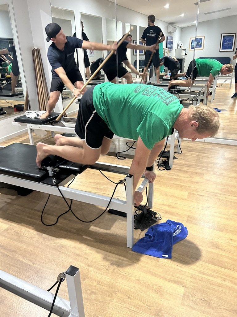 A man in a green shirt and black shorts performing an exercise on a reformer machine in a fitness studio, with other people exercising and mirrors on the wall.
