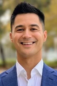 A smiling man in a blue suit jacket and white dress shirt outdoors with blurred trees in the background.