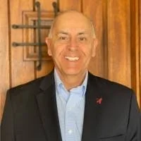 A smiling bald man in a suit and light blue shirt standing indoors with wooden walls and decor in the background.