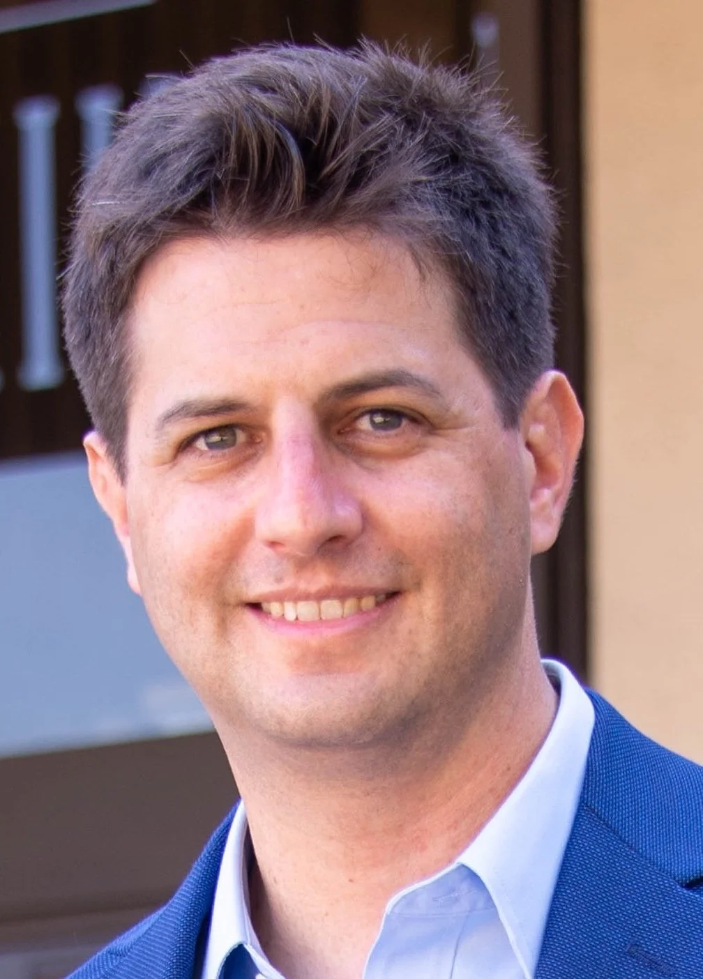 A man with short brown hair, blue eyes, and light skin, smiling, dressed in a white shirt and a blue blazer, standing outdoors in front of a house with a brown door.