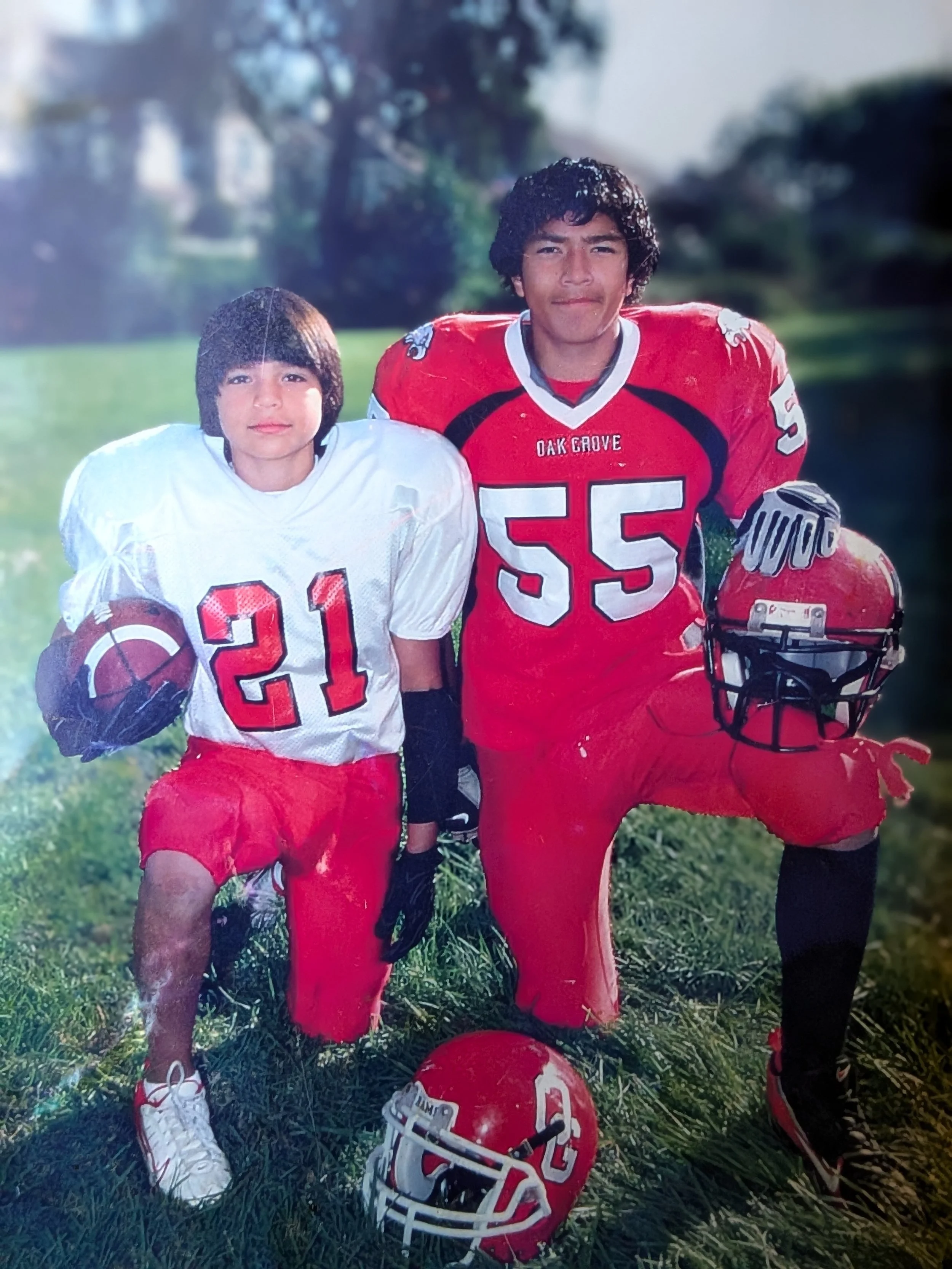 Two young boys in football uniforms, kneeling outdoors on grass. One boy wears a white jersey with the number 21, the other wears a red jersey with the number 55. They have football helmets nearby, with one helmet on the ground and the other in the younger boy's hand. The boy in red is holding a helmet with a logo. Both are smiling, with a background of trees and a blue sky.