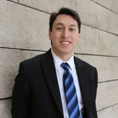 Young man in black suit and blue striped tie smiling, standing against a stone wall.