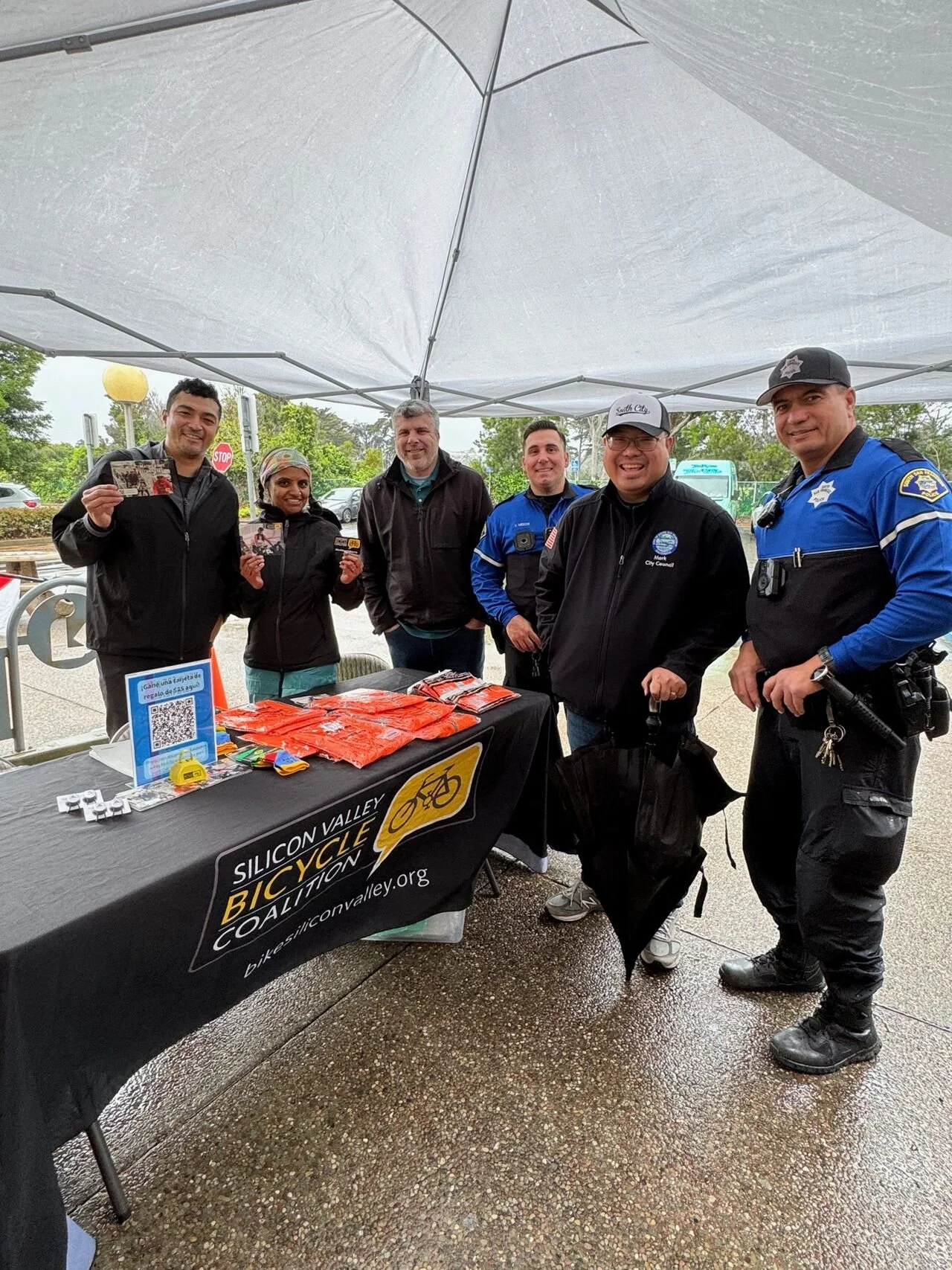 Group of six people standing under a canopy at an outdoor event, smiling, with a table displaying orange items and a banner for the Silicon Valley Bicycle Coalition.