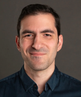 A professional headshot of a man with dark hair, wearing a dark collared shirt, smiling against a gray background.