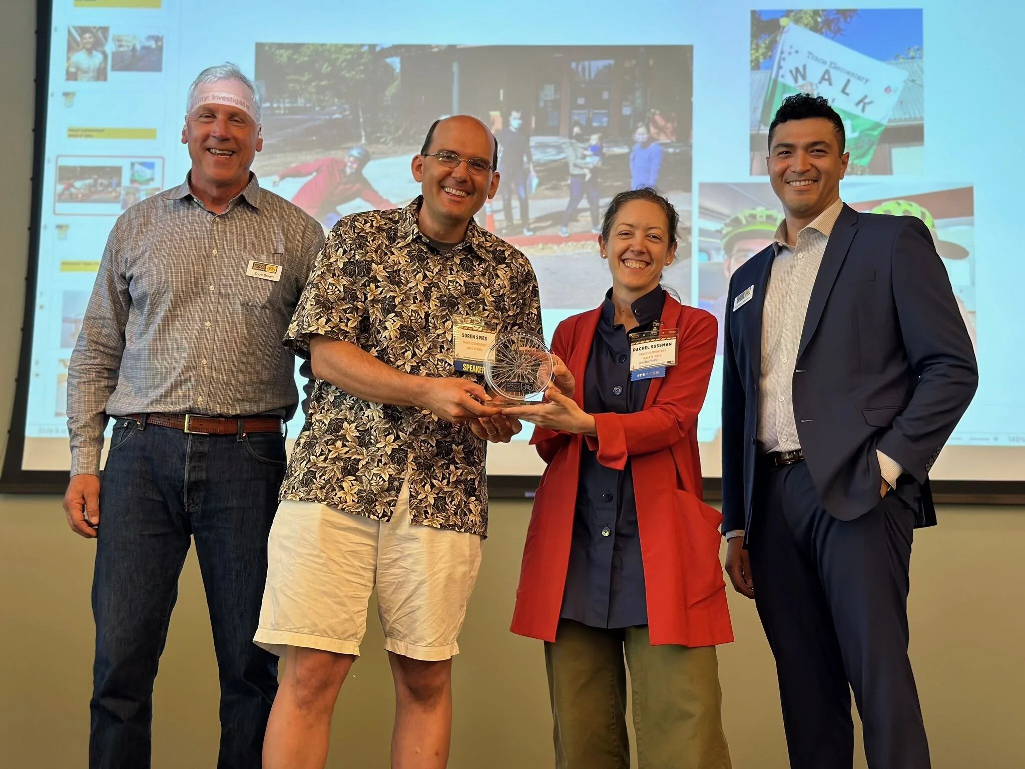 Four people standing in front of a presentation screen, smiling, with the two center individuals holding an award plaque. The background shows a projected slide with photos of people walking and a street sign that says ‘WALK’.